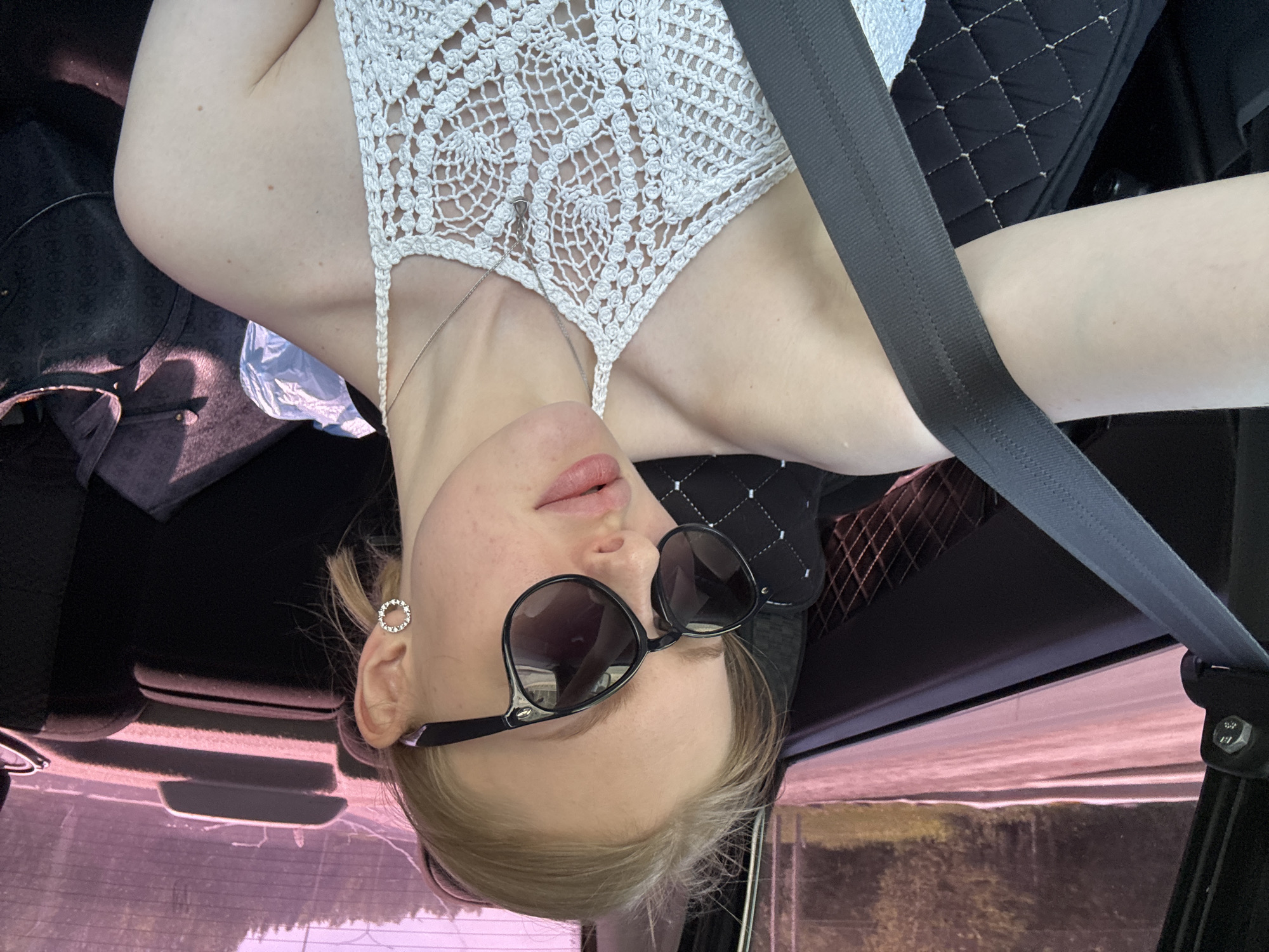 A young, fair-skinned woman is sitting in a car, wearing sunglasses and a white, crocheted top. A seatbelt is visible across her body. The background shows a view through the car window, with trees or foliage suggesting an outdoor setting. The image is taken at an unusual angle, possibly with the camera turned upside down. The location in Perm, Russia, is not directly discernible from the visual details of the photograph alone. The interior of the vehicle is visible, and also a handbag. The woman also wears a silver ear ring.