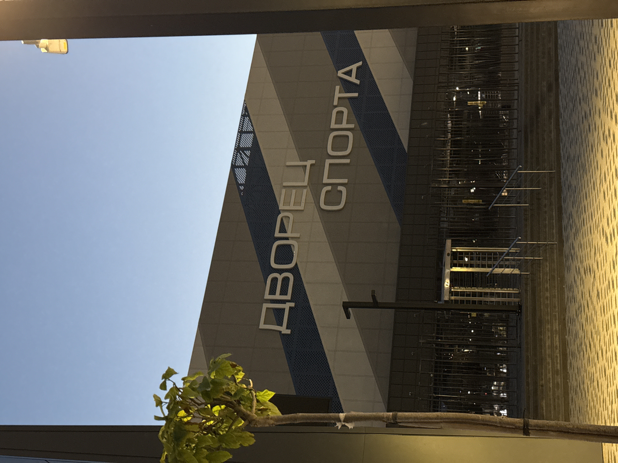 The image shows the "Sports Palace" in Rostov-on-Don, Russia. The building features a modern facade with diagonal blue and grey panels. Large white text on the building reads "ДВОРЕЦ СПОРТА" (Sports Palace). In front of the building, there is a metal fence and a turnstile entrance. A small tree with green leaves is visible on the left side of the frame. The ground in front of the steps is paved with light-colored tiles. The sky is a clear blue, suggesting it is daytime or early evening.