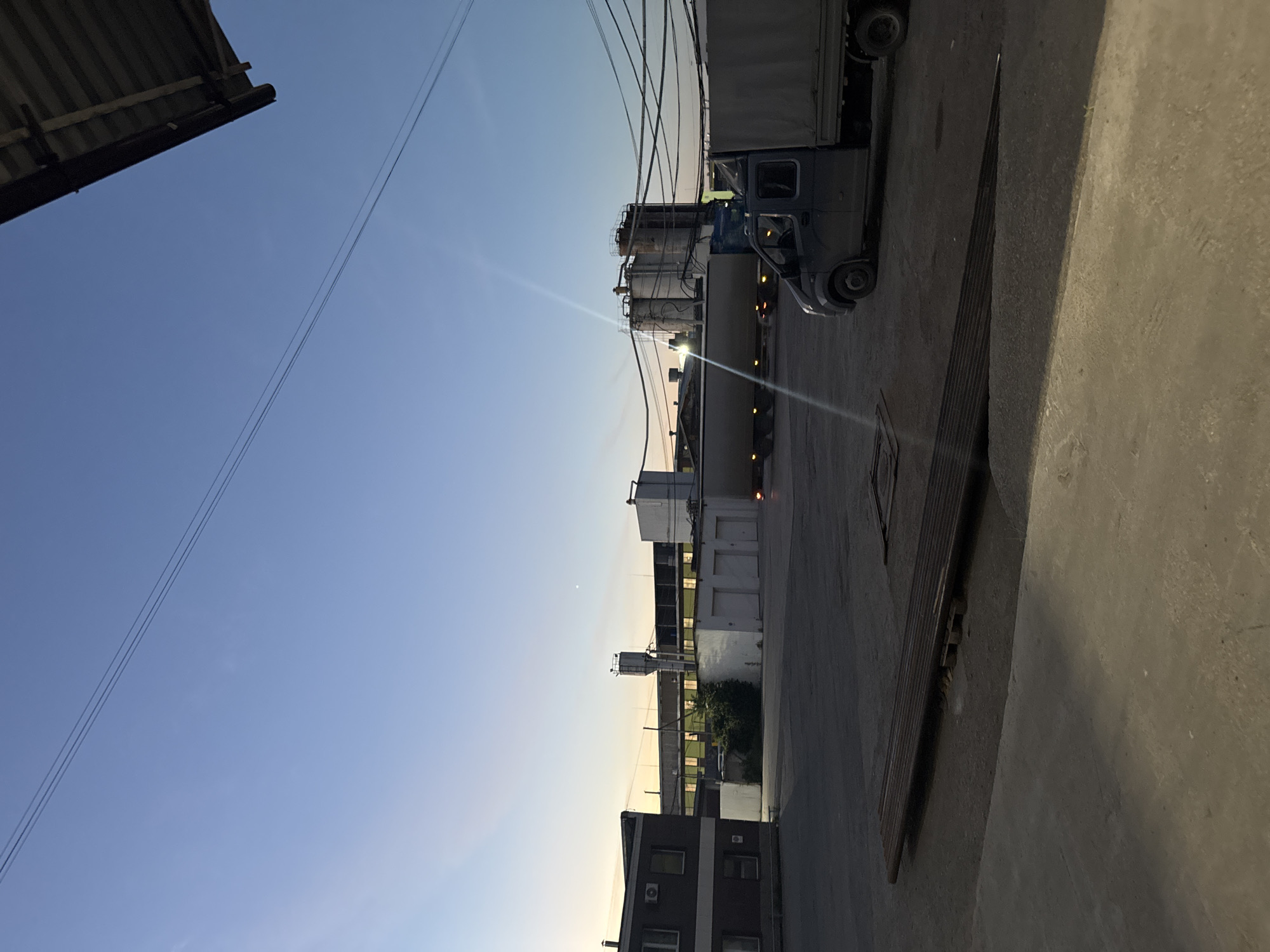 A wide shot captures an industrial area at dusk in Perm, Russia. The sky is a clear gradient of blue and pale yellow, with a few visible power lines crossing the frame. In the foreground, a concrete surface transitions to a darker asphalt lot. A long, flat object, possibly metal sheeting, rests on the asphalt.
Dominating the mid-ground are various industrial buildings. To the left, a multi-story building with dark-colored walls and visible windows, some with air conditioning units attached, stands out. Adjacent to it is a low-lying white building with several garage-style doors. Behind these structures, a taller, slender silo-like structure with a metallic top is visible. Further back, a long industrial building with many illuminated windows stretches across the scene. In the center-right, a large industrial tank complex with multiple cylindrical tanks is prominent.
Two trucks are present. A large tanker truck, its trailer largely obscured by the distance and lighting, is parked in the mid-ground, facing left. In the right mid-ground, a smaller, dark-colored truck with a covered bed is parked, facing left. No people are visible in the frame. The overall impression is one of a working industrial facility during the transition from day to night.
