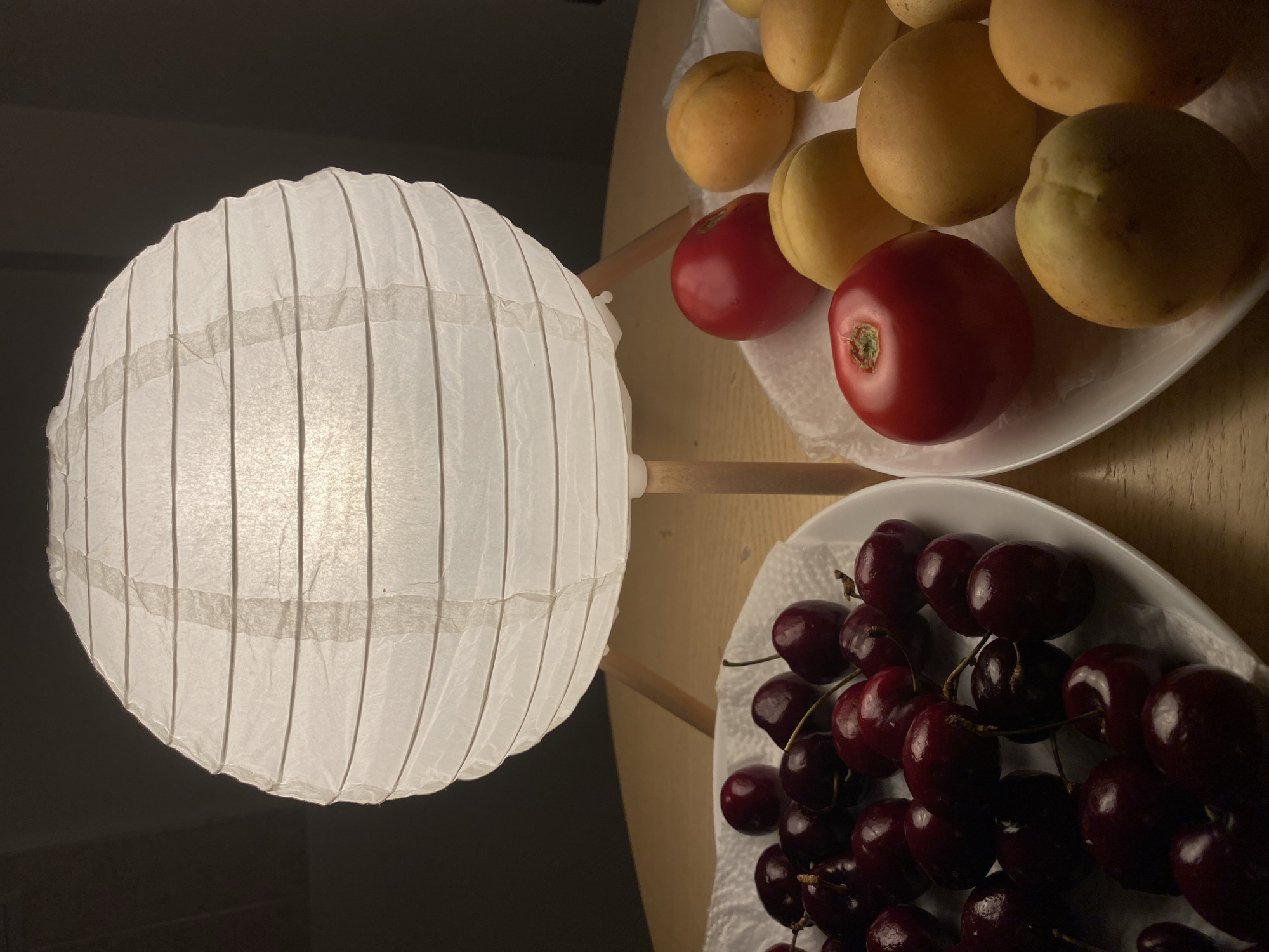 An overhead, close-up shot shows a white paper lantern on a wooden table. The lantern has horizontal pleats and is lit from within. Two white plates of fruit are arranged symmetrically on either side of the lantern. The plate on the left contains a pile of dark red cherries. The plate on the right contains a mixture of red tomatoes and yellow apricots. The background is dark and out of focus. The scene is well-lit by the lantern, casting soft shadows. The fruit appears fresh and is arranged casually on the plates.