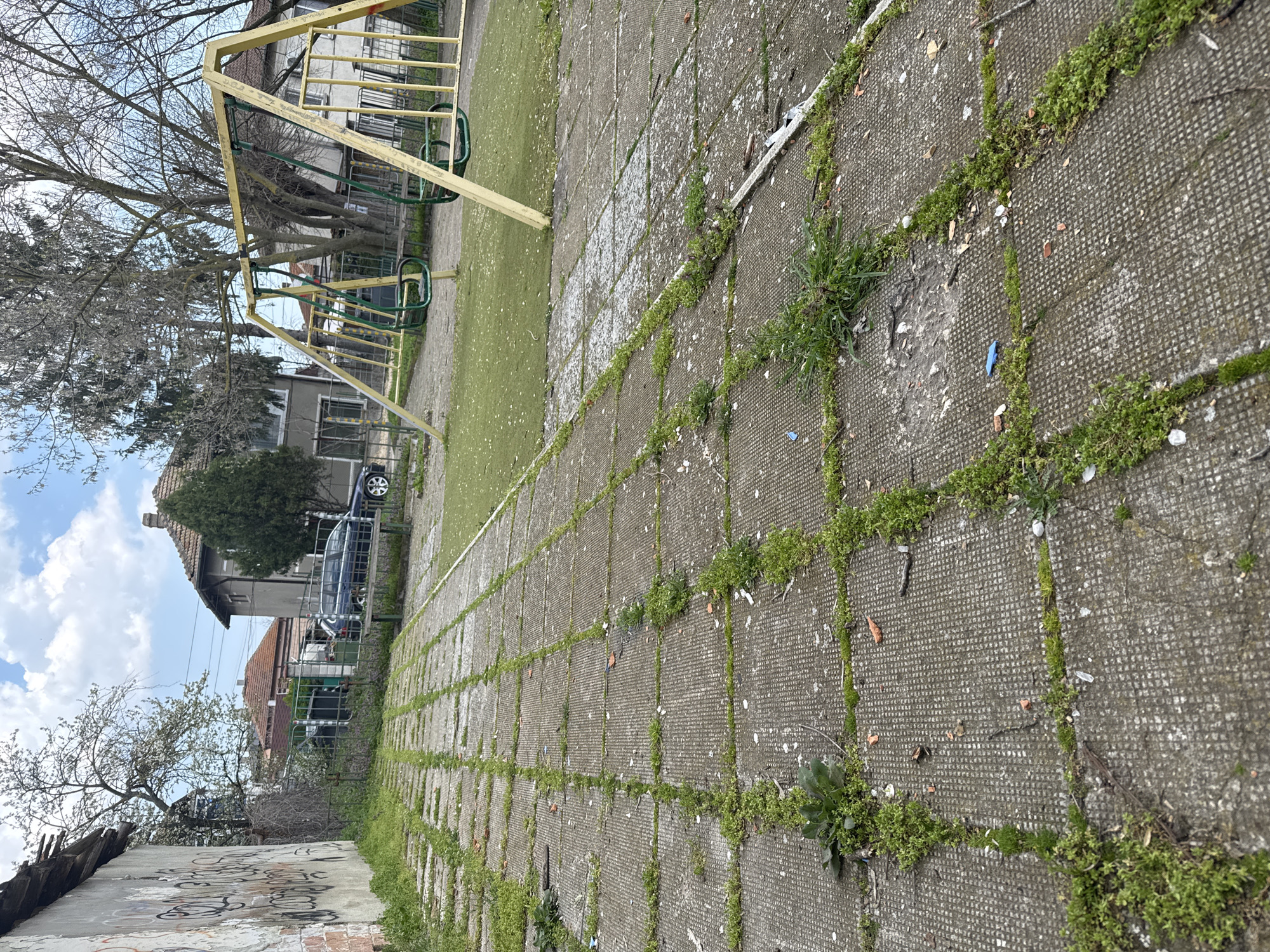 The image shows an overgrown, neglected playground in Topoli, Bulgaria. The ground is paved with square concrete tiles, heavily interspersed with moss and weeds growing in the cracks. A faded yellow swing set stands on a patch of artificial turf. In the background, there are residential buildings and a dark colored car parked nearby. The scene appears weathered and rundown, indicating a lack of maintenance. A graffitied, damaged wall is visible in the foreground. The sky is partly cloudy.