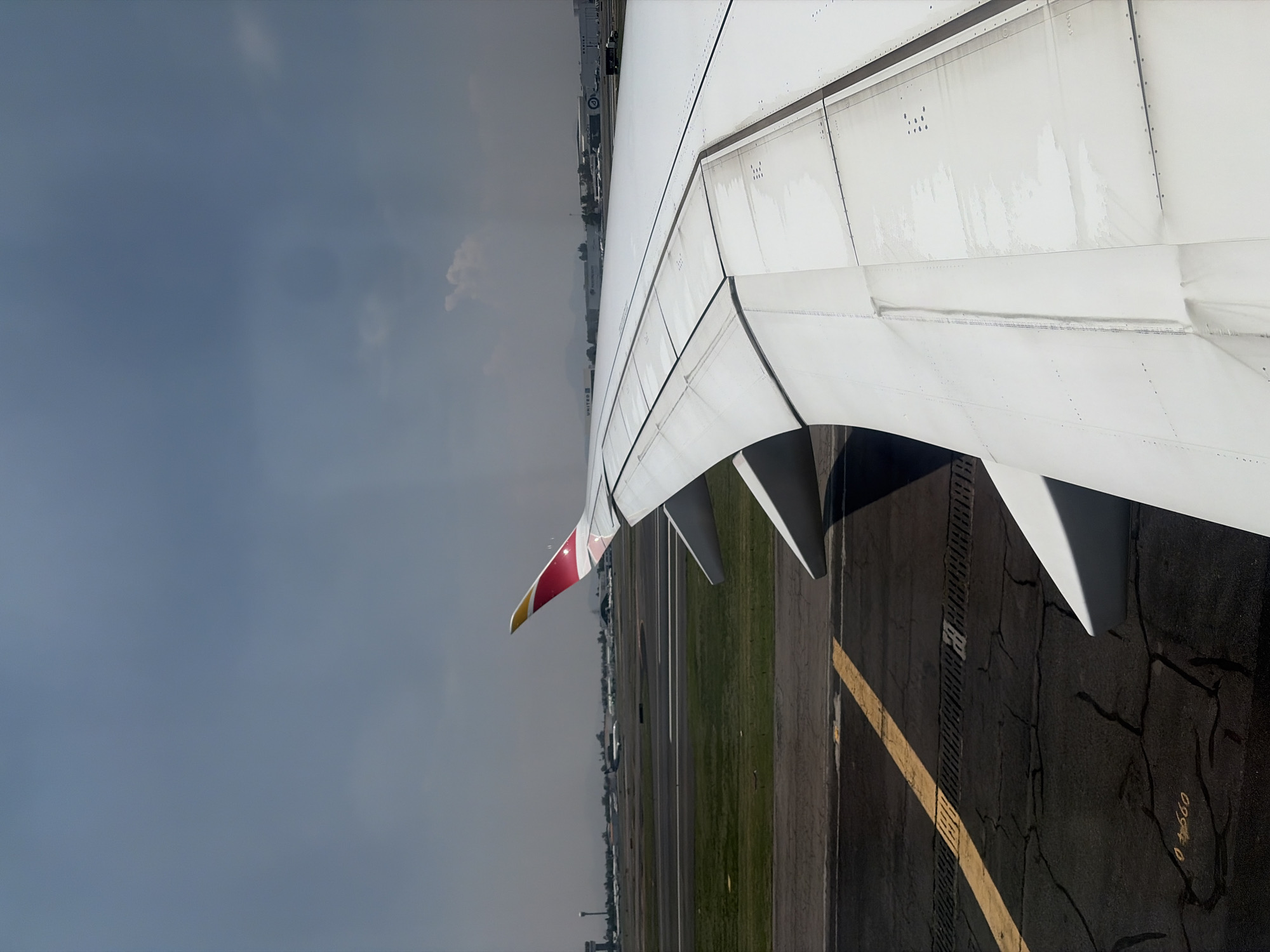 Here is a description of the image:
An aerial shot from inside an airplane shows part of the plane's wing and flaps extended over an airport runway. The wing is mostly white with a red and yellow wingtip. Below, the runway is made of cracked black asphalt with a clear yellow line running along it. There are grassy areas and buildings along the side of the runway. A gray, cloudy sky fills the other half of the frame.