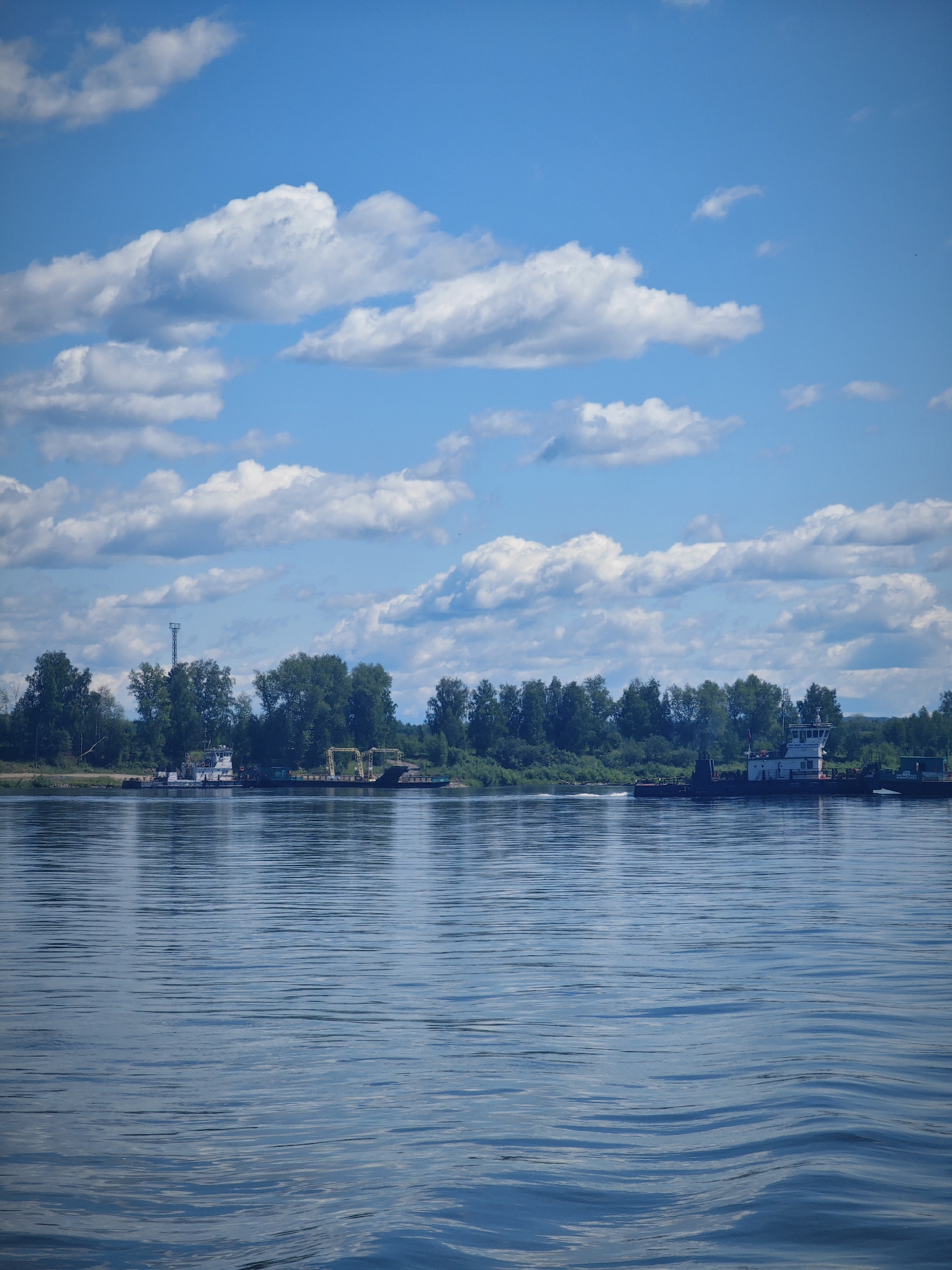 The image captures a wide, calm river on a bright, partly cloudy day. The water's surface is rippled, reflecting the sky and the lush green treeline on the opposite bank. Several boats are visible docked along the shore. One boat, a white tugboat, appears to be maneuvering, creating a wake. Another, larger vessel, possibly a barge or a ferry, is also present. A tall, lattice tower stands out against the trees on the left side of the frame. The sky is a vibrant blue, dotted with fluffy white clouds. The scene suggests a tranquil summer day at a riverside location, possibly near Krutoi Log, Russia, given the context.
