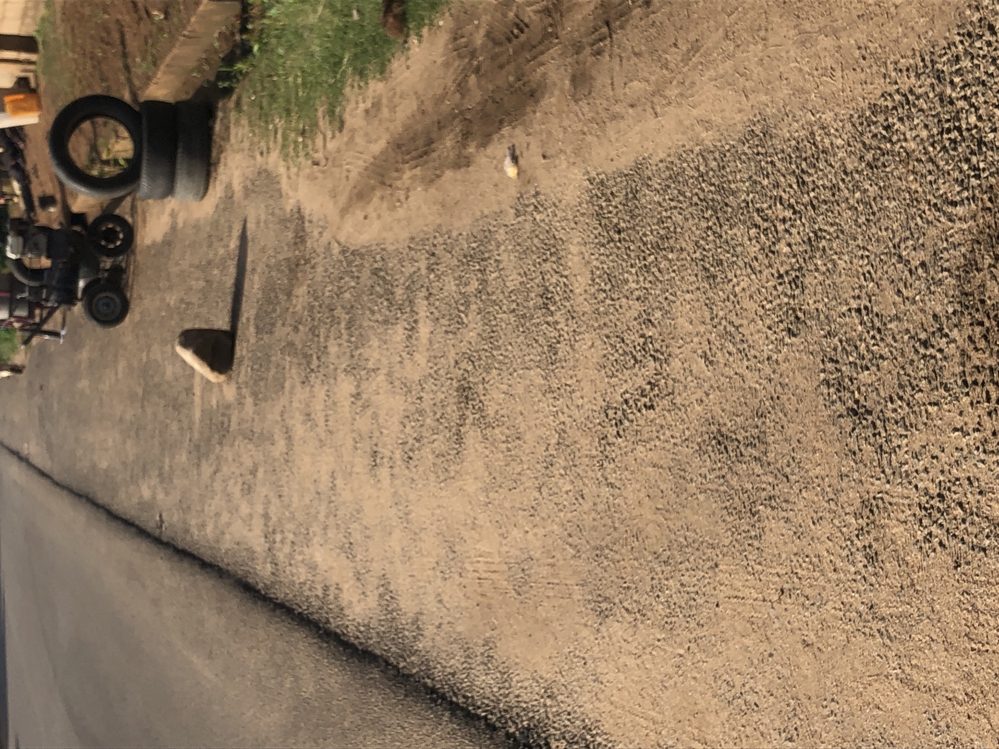 An upward-angle, medium shot captures a roadside scene with a rock in the foreground and construction equipment and stacked tires in the background. The ground is covered with a mix of gravel and dirt, with a paved road to the left. The rock, roughly oval in shape, is positioned to the left of center. Behind the rock, construction equipment with wheels and a metal frame is visible, alongside three black tires stacked one on top of another. A thin strip of green grass runs along the right edge of the frame, bordering a raised earthen berm. The lighting suggests it is daytime, possibly late afternoon, given the shadows cast. The location is identified as Unknown, Unknown.