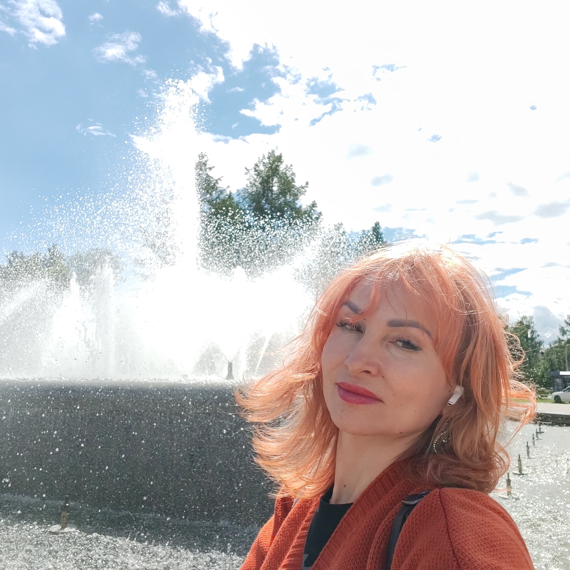 A woman is taking a selfie in front of a fountain on a sunny day. The fountain is spraying water high into the air and is located in the city of Chaikovsky, Russia. The woman has red hair and is wearing an orange cardigan and black top. She has an earbud in her ear and is looking at the camera. In the background, there are trees, clouds, and the sky.