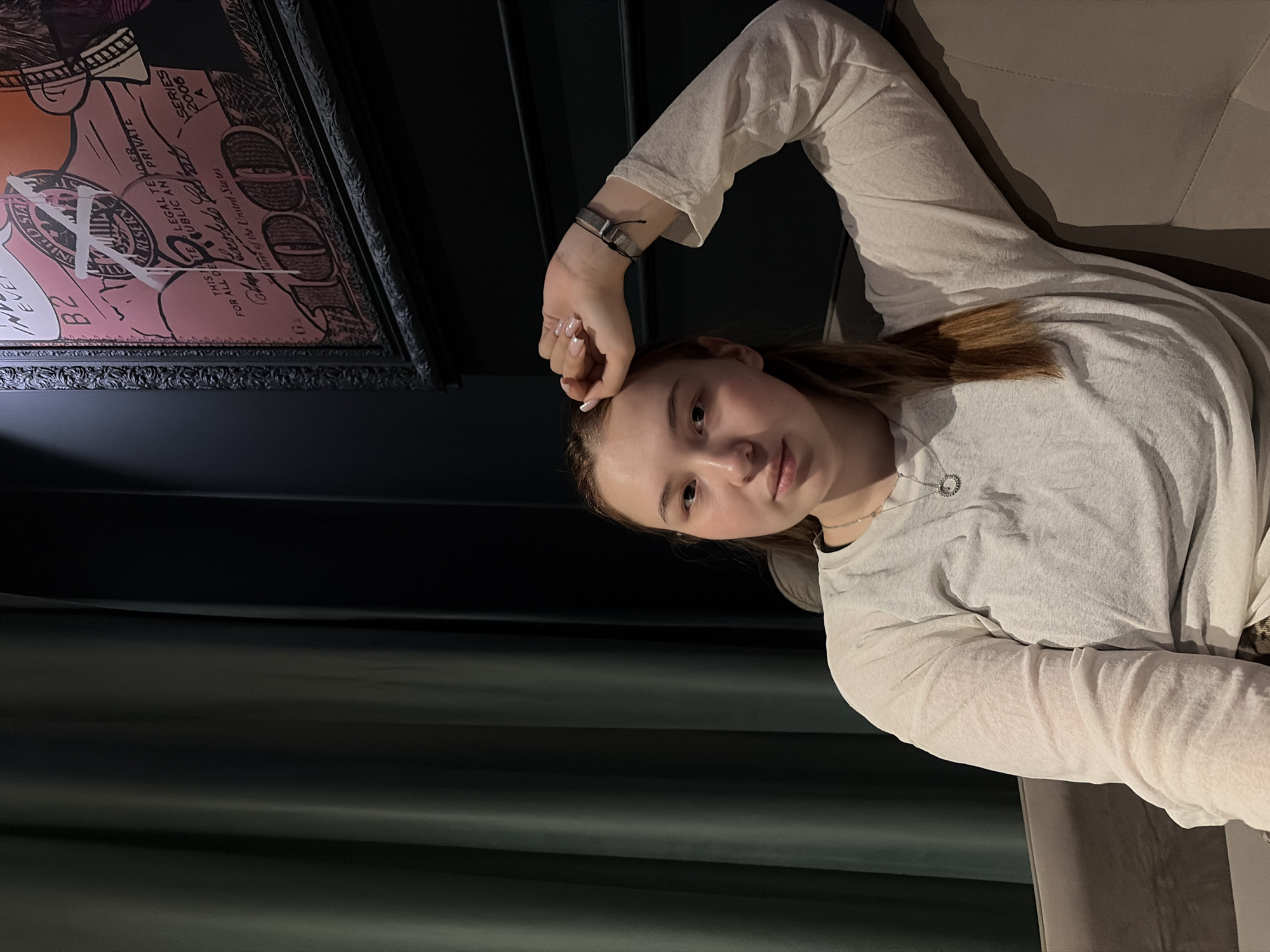 A young woman lies reclined, with a framed poster of a hundred-dollar bill visible on the wall behind her. The setting suggests an interior, perhaps a lounge or living room. The scene was captured at an undisclosed location within the city of Chaikovsky, Russia.
