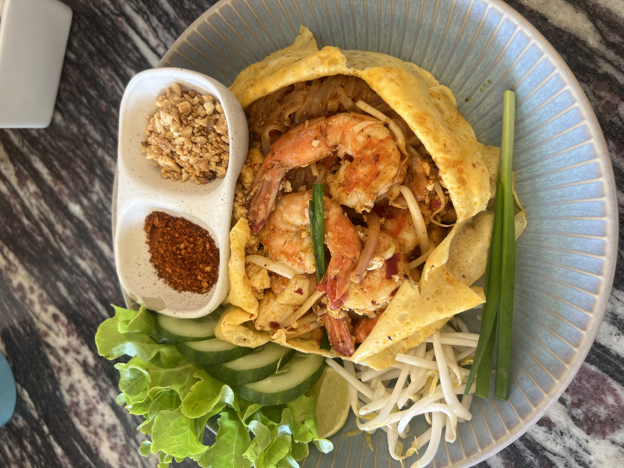A close-up, overhead shot shows a plate of Pad Thai served on a light blue, ribbed plate with a marble-patterned surface. The Pad Thai is presented with a thin omelet draped over the noodles, and garnished with large shrimp, bean sprouts, sliced cucumber, lettuce leaves, a lime wedge, chopped peanuts, and chili flakes served in a divided ceramic dish. Two green onions are placed on the side of the plate. The lighting suggests it is daytime, with the food illuminated from above. The location is identified as Koh Phangan, Thailand.