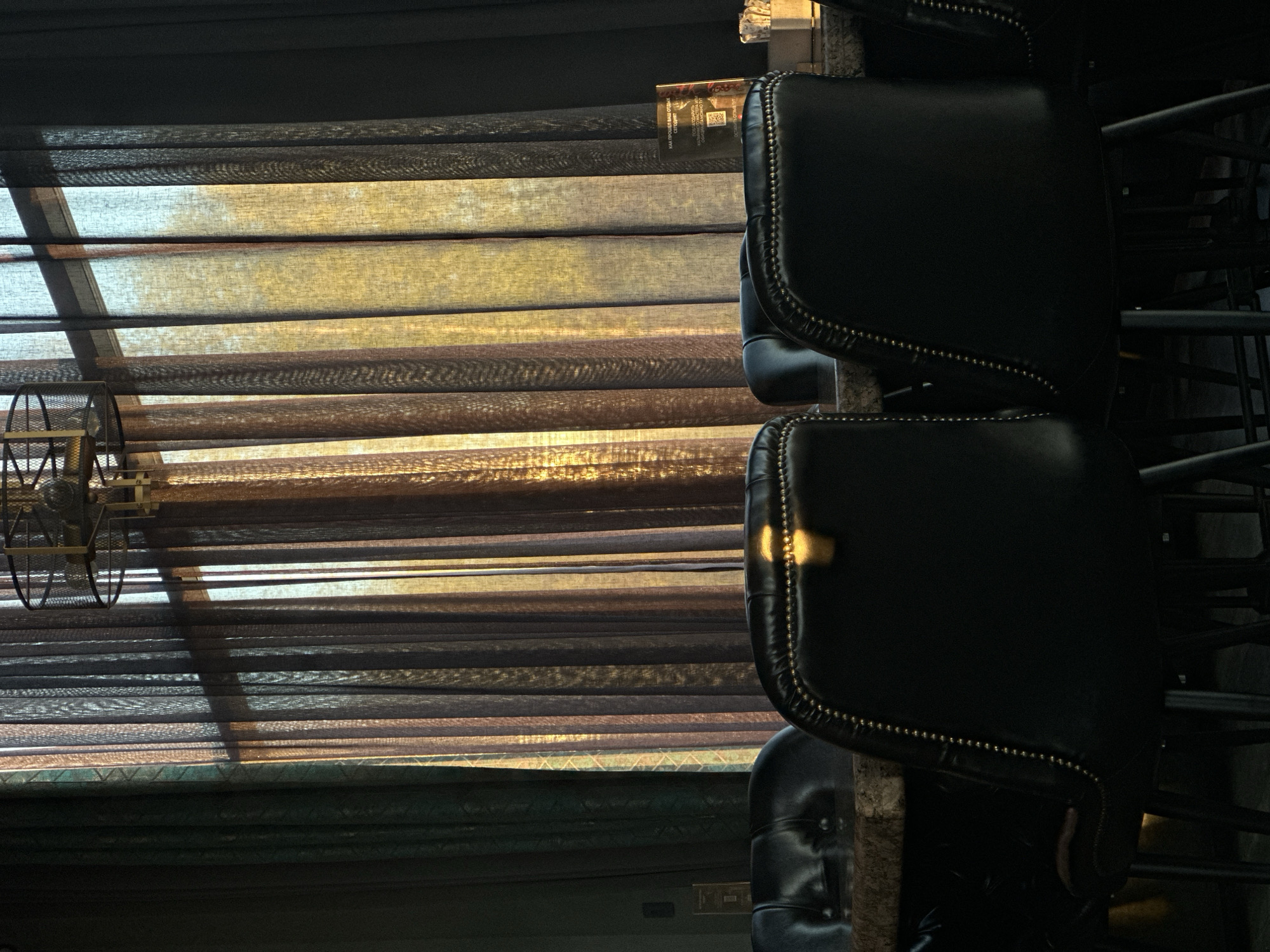 The image depicts a stack of dark leather chairs with nailhead trim to the right of a window covered with horizontal blinds. The blinds are partially open, allowing sunlight to stream through, creating patterns of light and shadow. A metal fan is visible in the lower left corner, partially obscured by the blinds. The overall lighting is dim, with the sunlight providing a focal point of brightness. The location is not identifiable as Votkinsk, Russia, based on the visible elements.