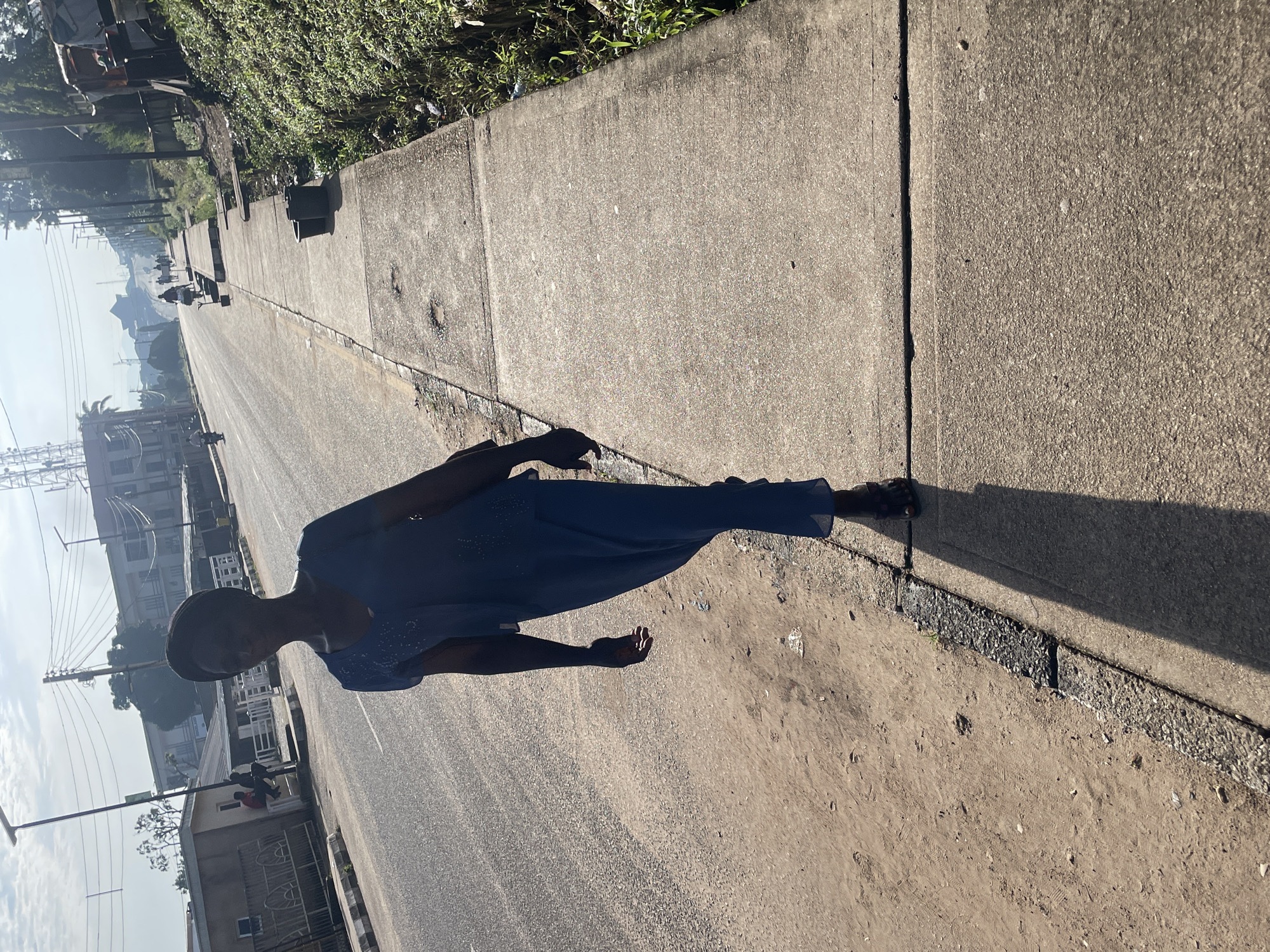 A full-body, low-angle shot shows a dark-skinned person walking on a sidewalk next to a road. The person is wearing a dark blue, long-sleeved dress with a matching headscarf. Their shadow stretches out before them on the sidewalk. The sidewalk is cracked and uneven, with patches of dirt and debris. The road is paved and appears to be a busy street, with vehicles and other pedestrians visible in the distance. The background includes buildings, power lines, and lush green vegetation. The sky is a bright, hazy blue. The scene is lit by bright sunlight, casting strong shadows.