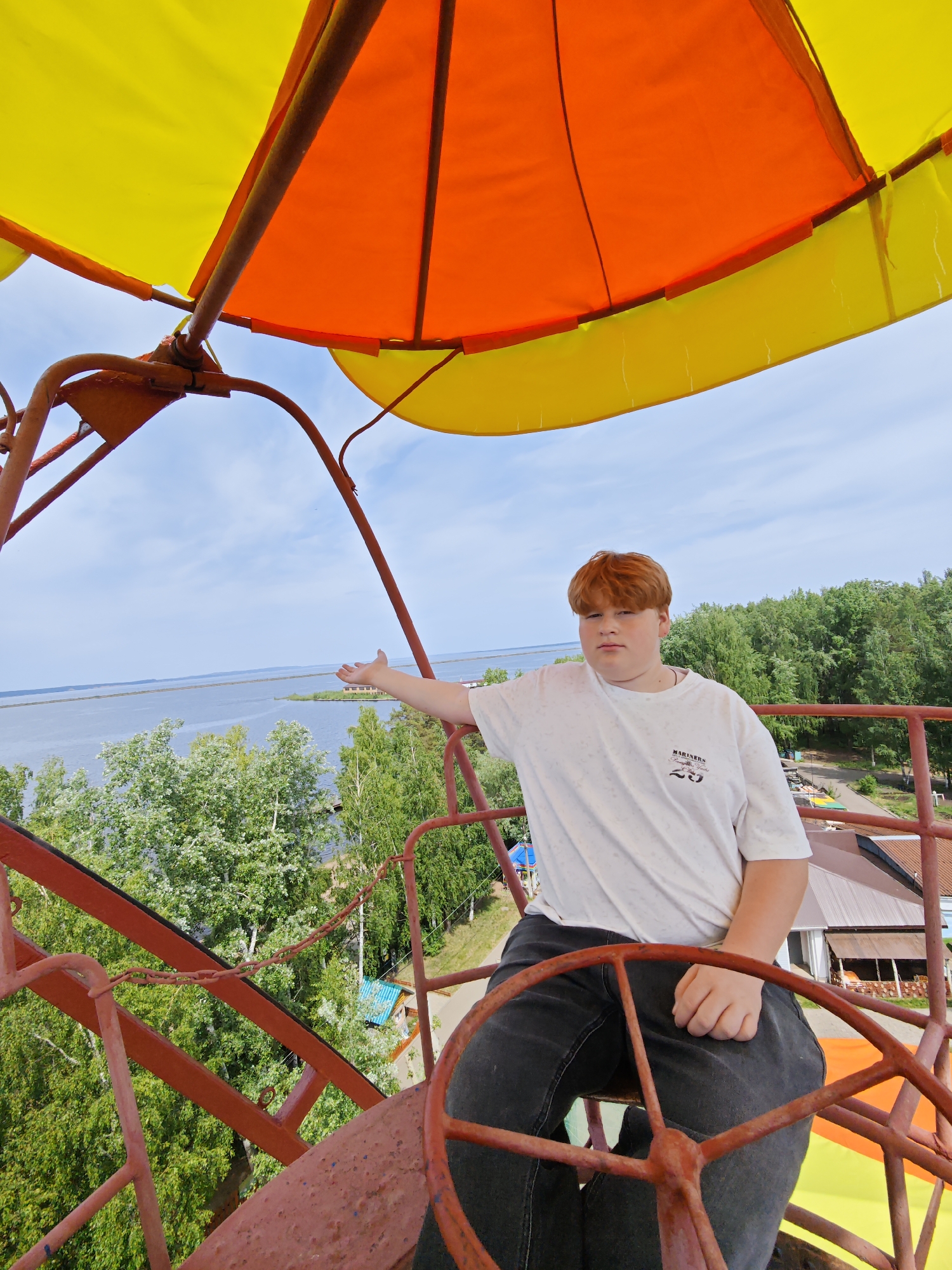 A full-length shot depicts a young, fair-skinned male with red hair seated within a red metal structure under a yellow and orange canopy. He is wearing a white t-shirt with black print and black jeans. His left arm is outstretched. The setting includes a body of water visible in the background, along with dense green trees and vegetation. A building with a brown roof is partially visible. The scene is located in the city of Chaikovsky, Russia.