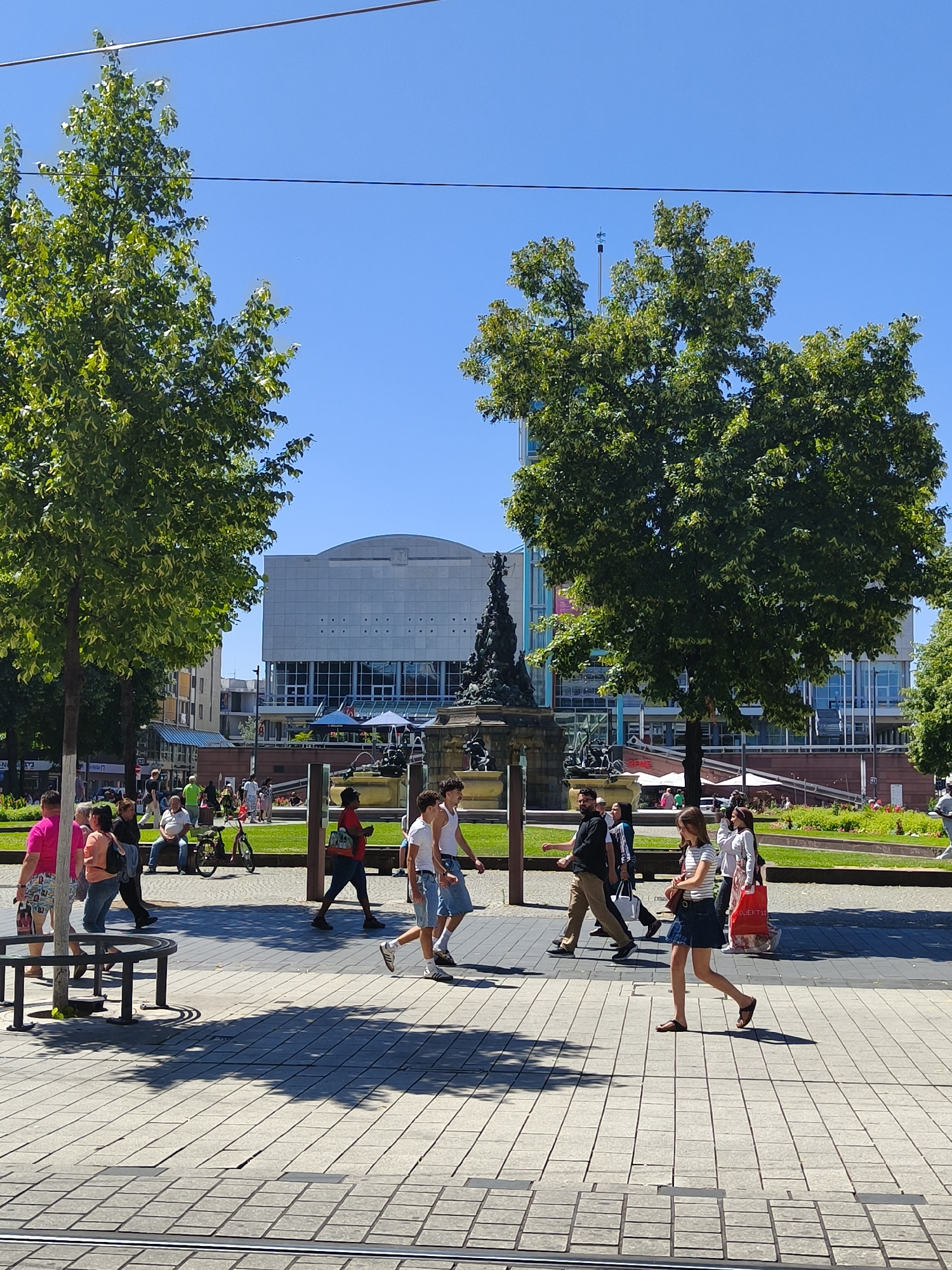 This is a photograph of the Paradeplatz, Mannheim, Germany, on a sunny day. Several people are walking across a paved plaza, some are sitting on benches, and a bicycle is parked on the side. In the background, there is a large modern building with a white facade and a sculpture with a fountain. Two large trees with green leaves are in the foreground, casting shadows on the plaza. The sky is clear and blue.