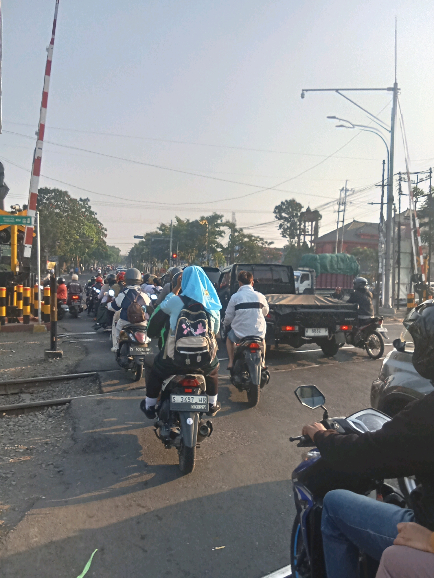 The image shows a traffic scene in Kaliwungu, Indonesia, with a railway crossing in the foreground. A long line of motorcycles and a truck are stopped, presumably waiting for a train to pass. People are riding on the motorcycles, some wearing helmets and others with headscarves. The environment appears to be urban, with trees and utility poles visible in the background. The sky is clear and the sun is shining, casting shadows on the road. The image captures a moment of congestion on a typical day in this Indonesian city.