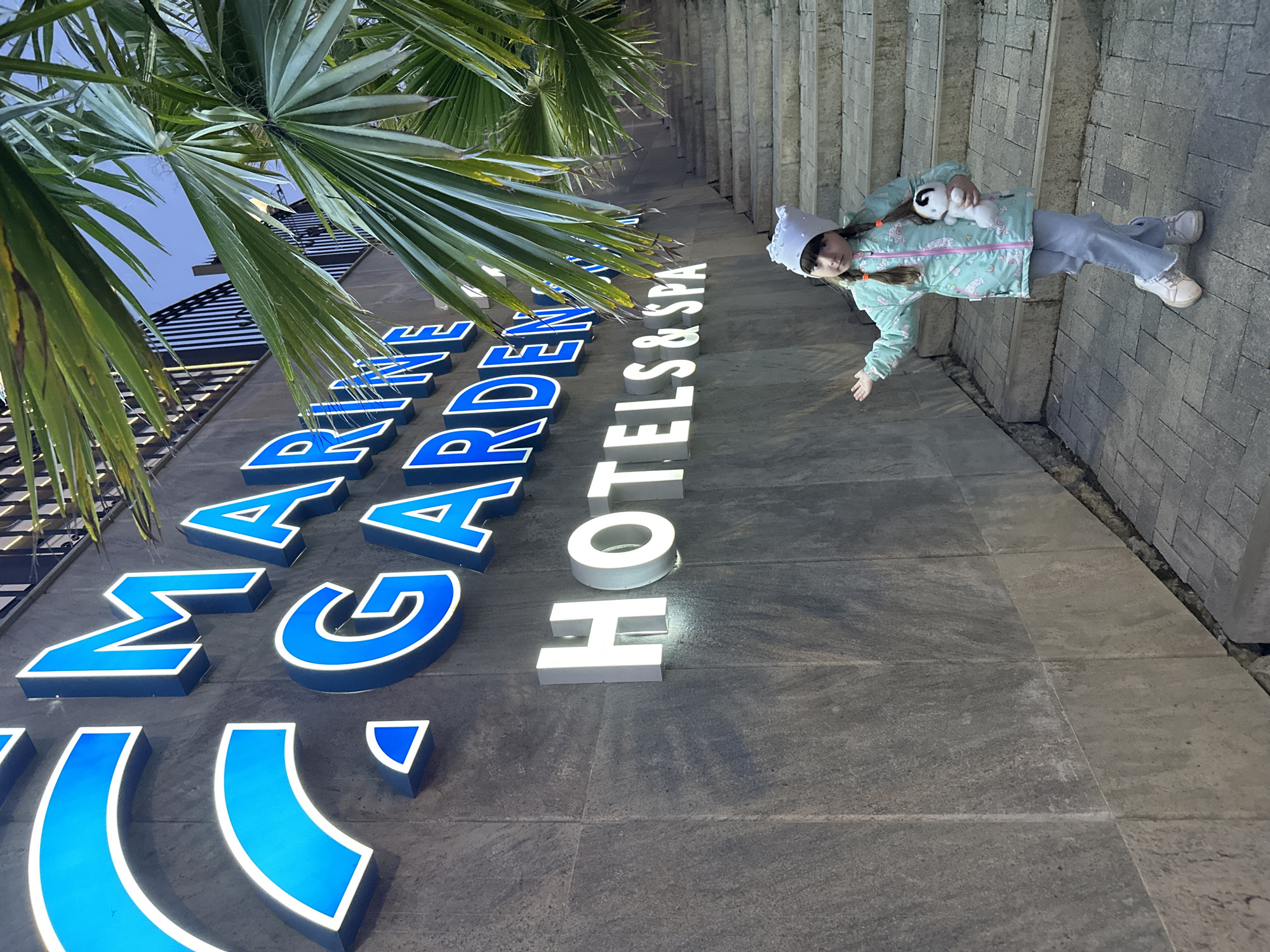 An evening view in Sochi, Russia, captures a young girl standing beside a large, illuminated hotel sign. The sign is vertically mounted on a dark grey stone wall, displaying the text "MARINE GARDEN HOTELS & SPA." The letters for "MARINE GARDEN" are bold blue with white illuminated edges, while "HOTELS & SPA" are white and brightly lit. Lush green palm fronds partially frame the top and left of the scene, suggesting a warm climate.
The girl, facing the camera, wears a light blue and white patterned jacket, light grey trousers, a white knitted hat, and white sneakers. Her hair is in two braids. She holds a black and white stuffed panda toy in her left arm and has her right arm slightly extended. The ground is paved with grey and lighter grey rectangular tiles. The illumination of the sign indicates it is dark outside, likely evening or night. The girl's expression is neutral and calm.