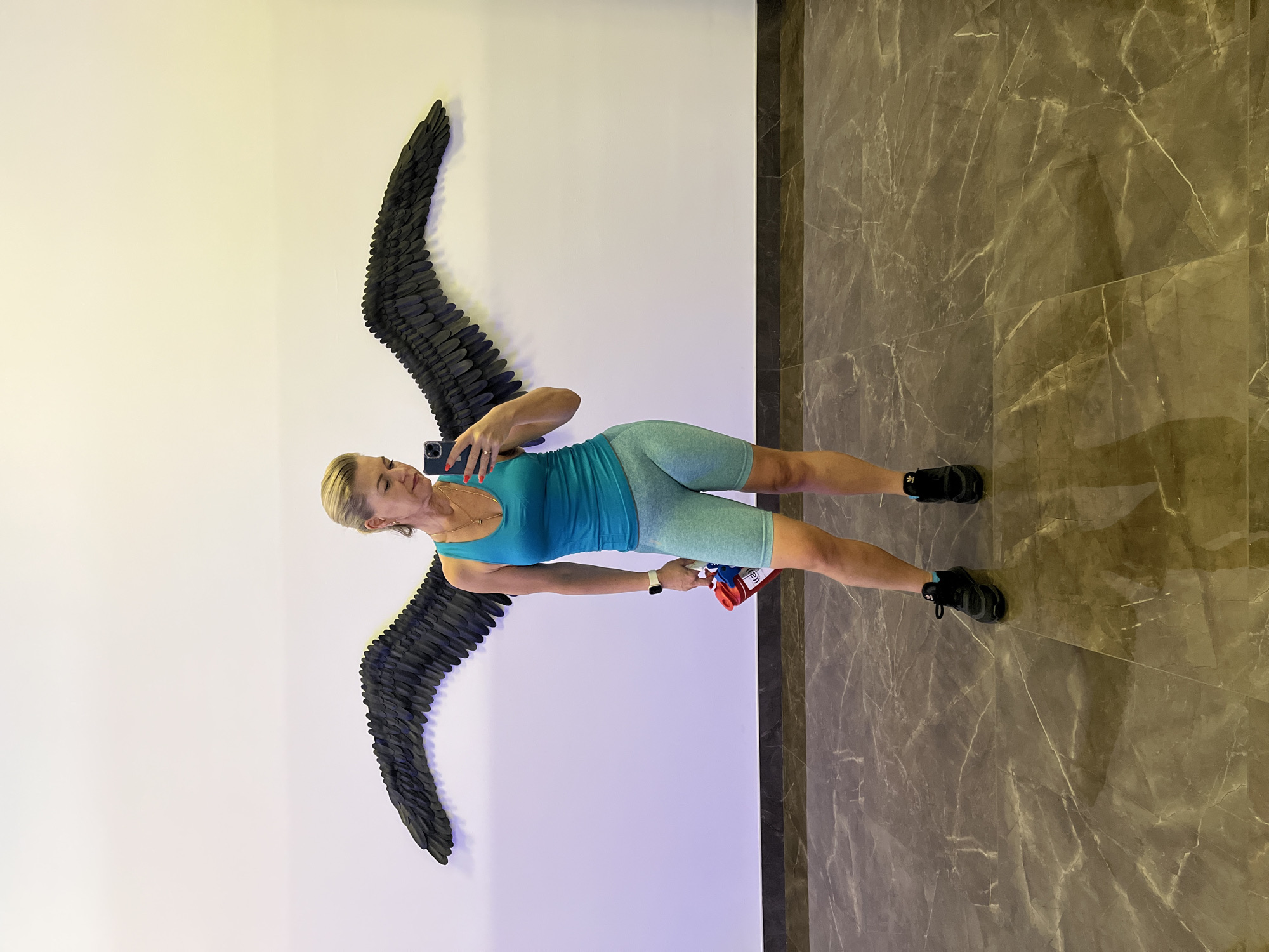 A blonde woman in athletic wear stands facing a wall with large black feathered wings mounted on it, taking a selfie with her phone. She is wearing a teal tank top and light blue cycling shorts, along with black sneakers. In her left hand, she holds a red and white object, possibly a water bottle or a small cooler. The wall behind her is white and illuminated with purple light on the left side. The floor is a dark, polished marble with a vein pattern. The overall setting appears to be indoors, possibly a modern gym or a public space with artistic installations. The location of Kaliningrad, Russia is not discernible from the image itself.