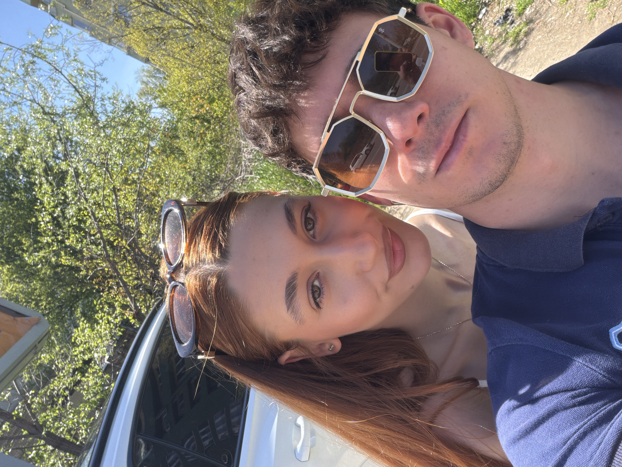 A selfie depicts a couple standing outdoors. The man, positioned at the top, wears sunglasses with a metallic frame. He has dark hair and a light complexion. The woman, located at the bottom of the frame, has long, reddish-brown hair, with sunglasses perched atop her head. She also has a light complexion. The background features trees and a bright sky. A white car is partially visible on the left side of the frame. Based on available details, determining the precise location in Sofia, Bulgaria is not possible.