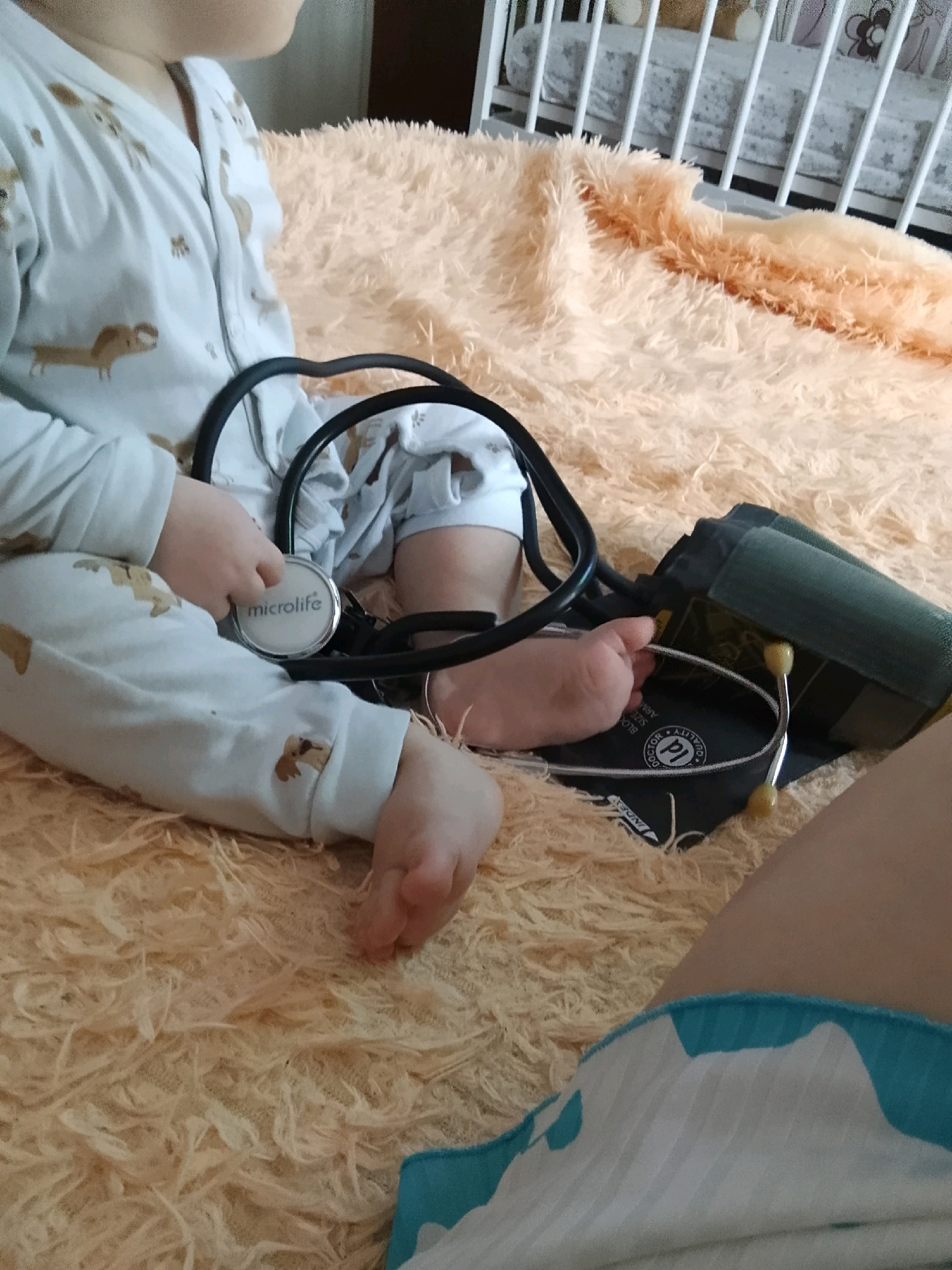 A close-up, low-angle shot shows the legs and torso of a toddler lying on a fuzzy, peach-colored blanket. The toddler is wearing light blue pajamas with a brown animal print. A black stethoscope is draped around the toddler's neck and chest, with the earpieces resting near their hands. To the right of the stethoscope, a blood pressure cuff lies on the blanket, with the stethoscope chestpiece resting on it. The background reveals a white crib with bars and some bedding. The scene appears to be indoors, on a bed or crib, with no specific city or location identified within the frame.