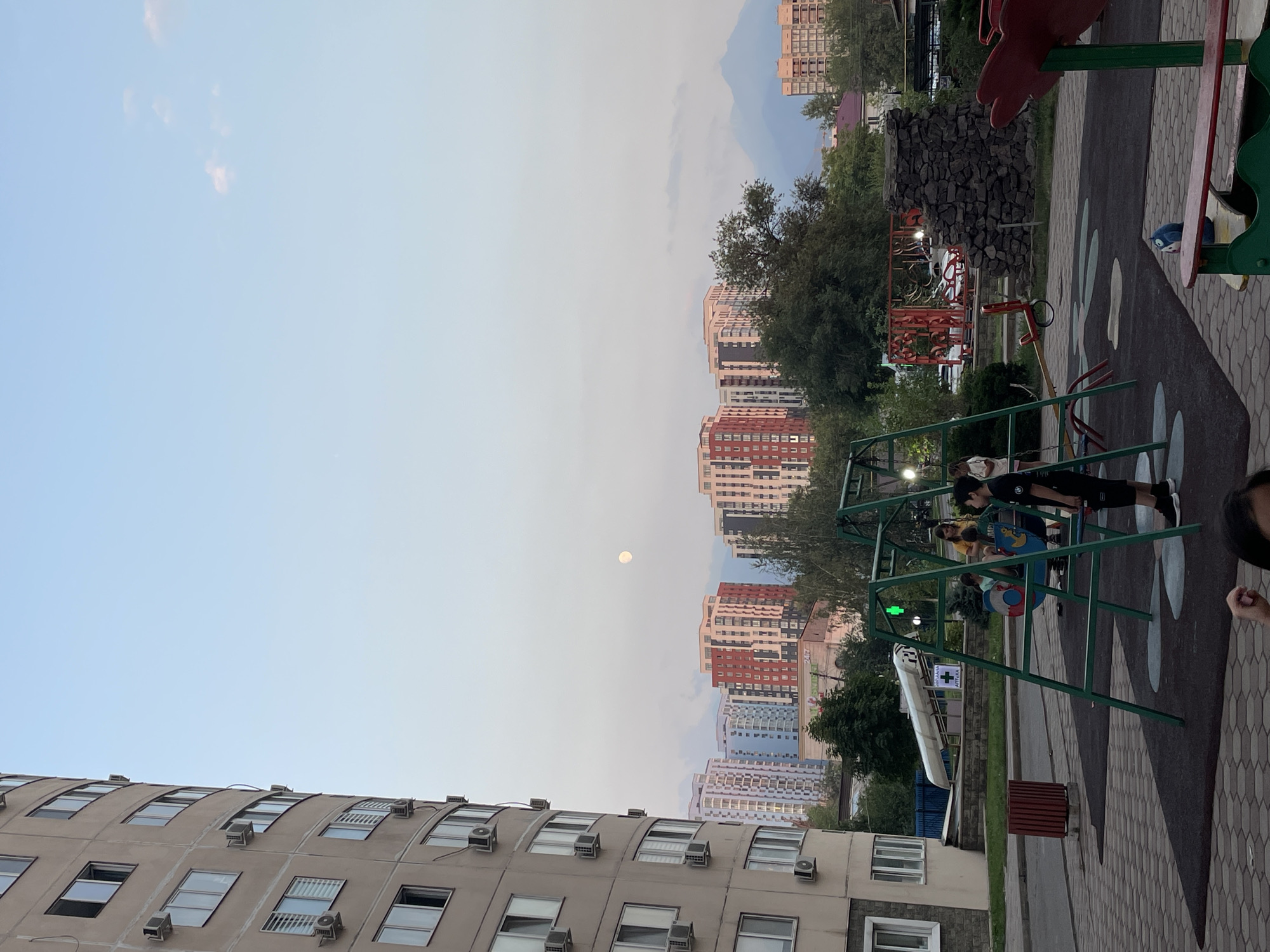 The image captures a residential courtyard in Almaty, Kazakhstan, during what appears to be late afternoon or early evening. In the foreground, a children's playground features a green swing set with several children present. One boy in a black t-shirt and shorts stands near the swing, while other children are seated on the swings. A seesaw is also visible. To the left, a large, curved apartment building dominates the frame. In the mid-ground, a cluster of colorful, modern apartment buildings rises above mature trees. In the distance, hazy mountains are visible under a pale sky, with a full moon prominent in the upper sky. A red trash bin is situated on the paved area of the courtyard. The overall impression is one of urban residential living with recreational amenities.
