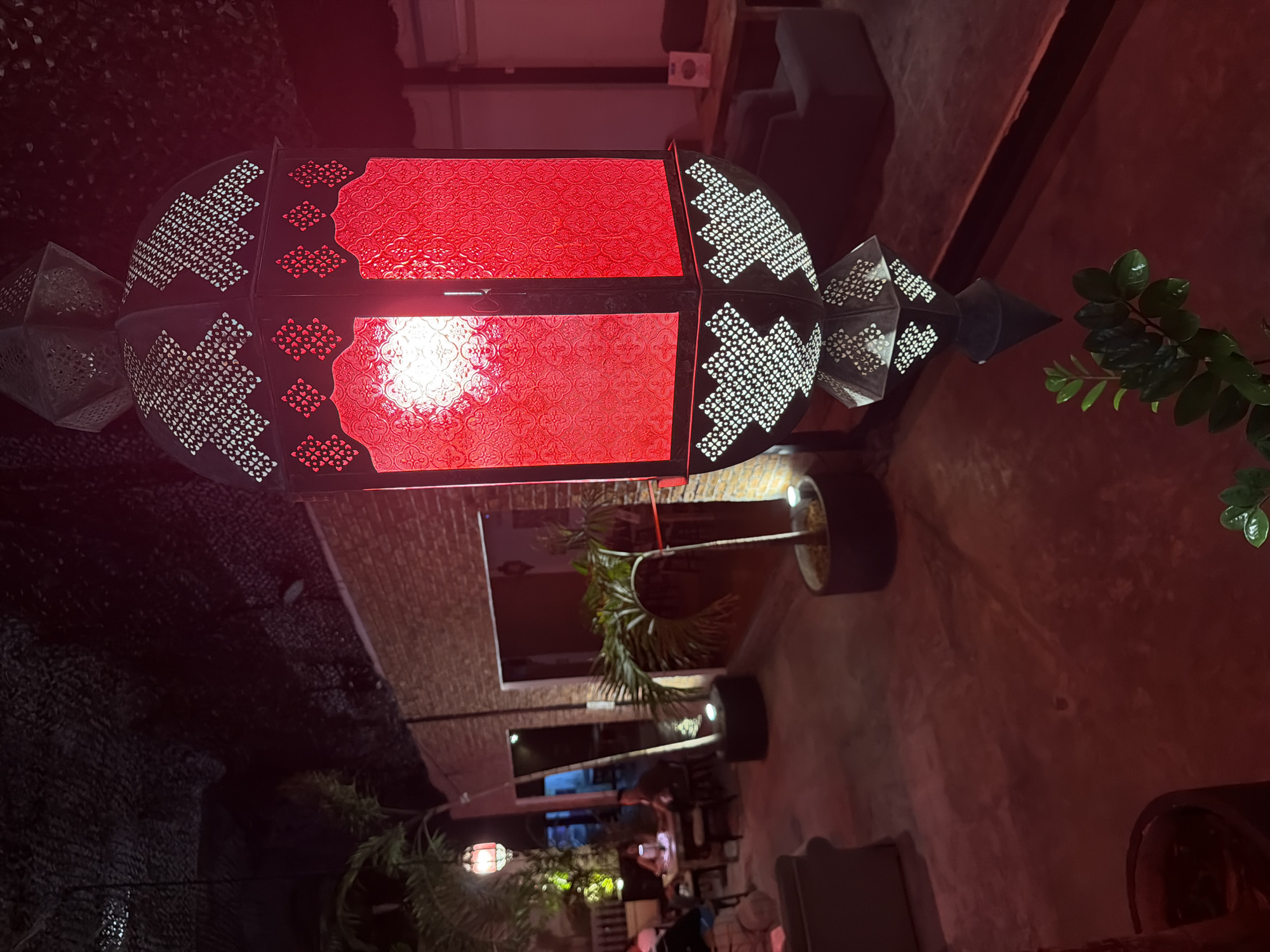The image shows an outdoor seating area at night. A large, ornate red lantern hangs prominently in the foreground, casting a warm glow. In the background, several people are seated at tables, some appearing to be dining. Lush green plants and brick walls contribute to the ambiance. The overall scene suggests a relaxed and inviting atmosphere, possibly a restaurant or bar. The location is identified as บ้านบางรักษ์, Thailand.