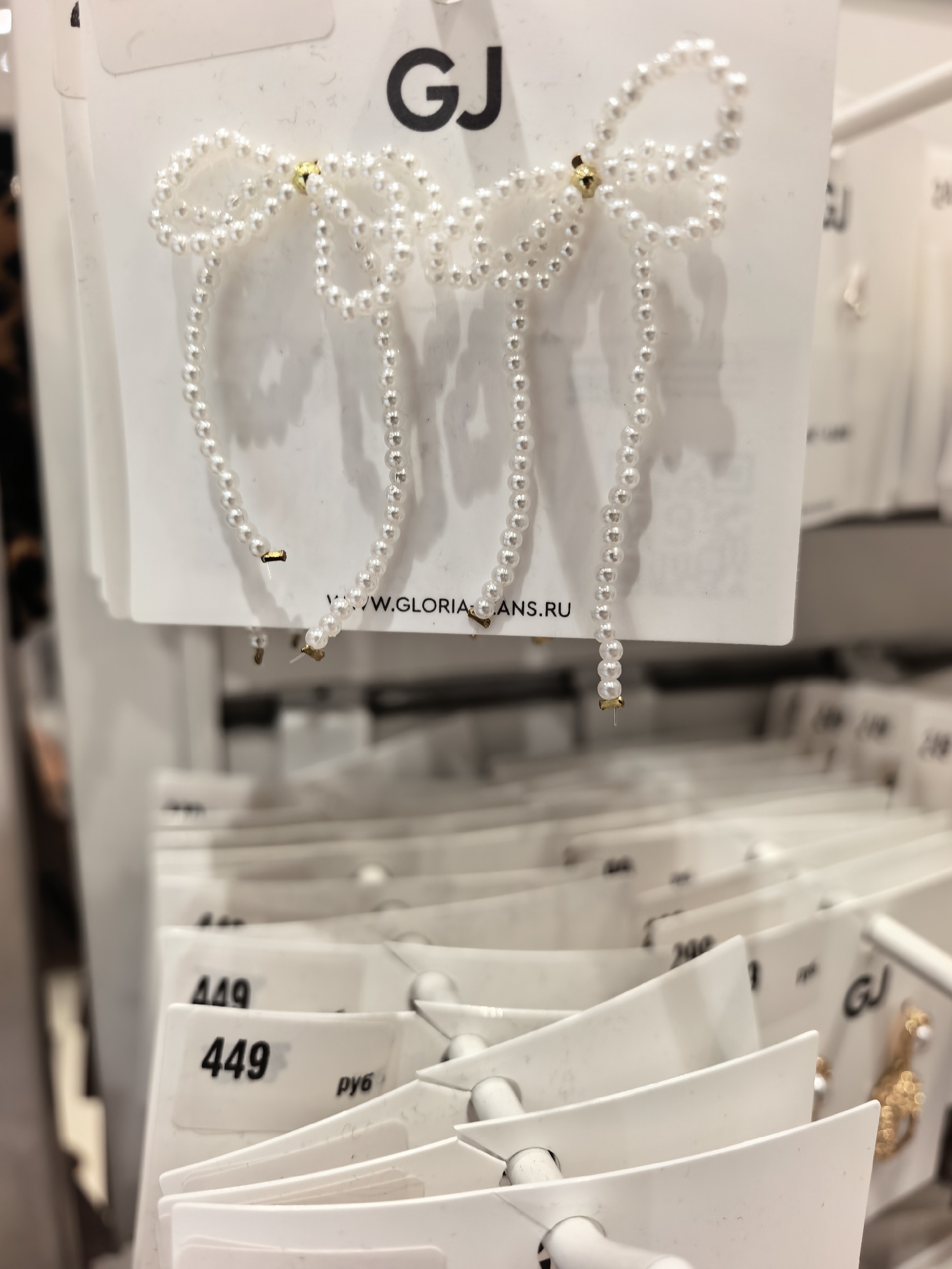 The image shows a display of pearl-like earrings shaped as bows. The earrings are attached to white cardboard tags with the brand "GJ" visible on one tag. Another tag shows a website address, "WWW.GLORIA-ANS.RU". In the foreground, there are other price tags, one of which reads "449 руб" (449 rubles). The items are displayed on a rack in what appears to be a retail store. The overall lighting suggests an indoor environment. This scene is consistent with a retail setting in Sterlitamak, Russia, where such fashion accessories are commonly sold. There are no people visible in the image.