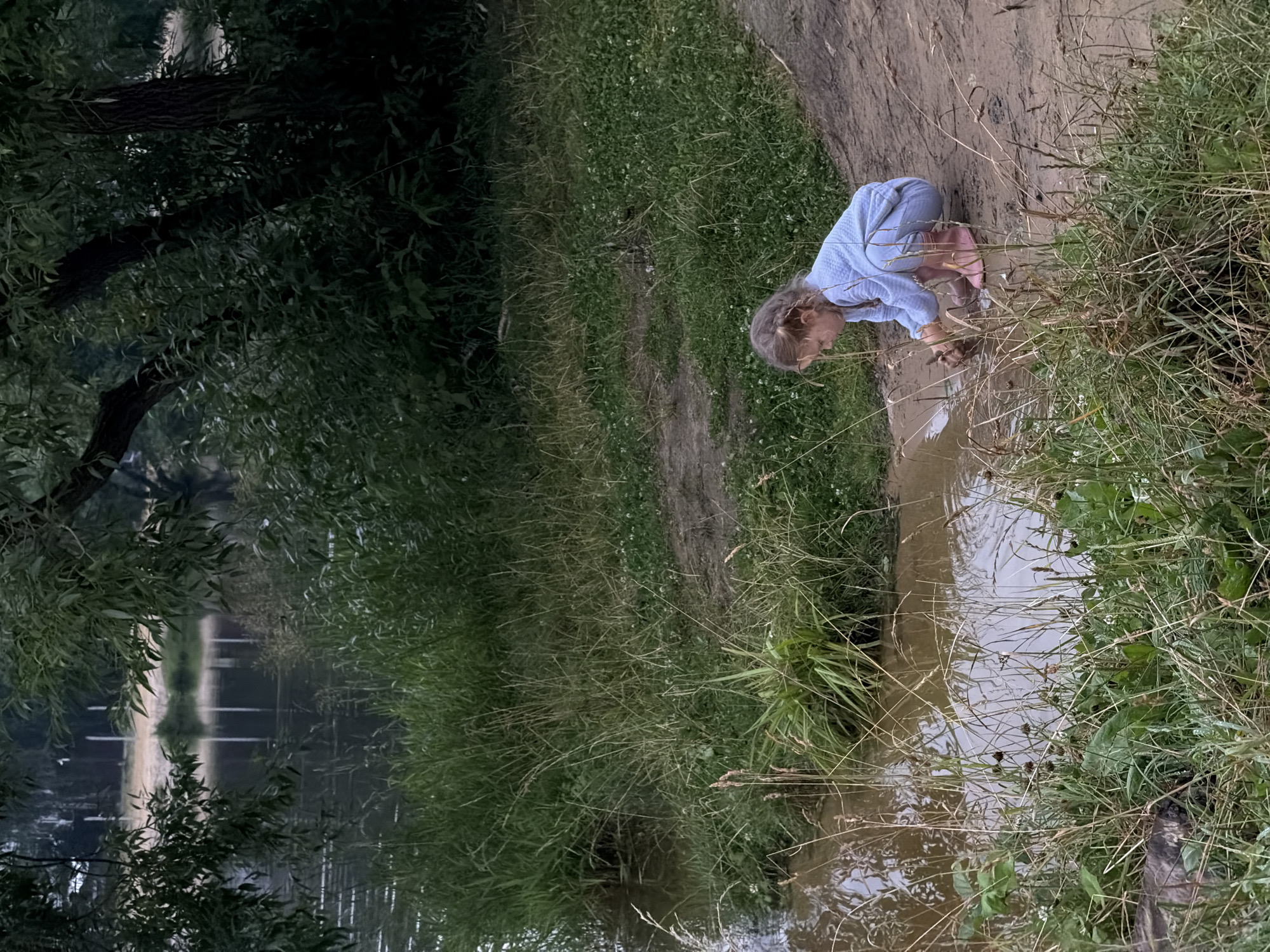 A young girl with blonde hair in a light blue quilted tracksuit and pink boots is crouching by the muddy bank of a body of water, playing with something in the shallow water and mud. She is positioned in the lower right quadrant of the frame, facing towards the left. The environment is natural, with lush green grass and reeds growing along the water's edge, and trees and foliage visible in the background. The water appears calm and reflects the overcast sky. The ground where the girl is playing is a mix of mud and dirt. The overall scene suggests a peaceful outdoor activity in a rural or semi-rural setting. This location is identified as Malakhovka, Russia.