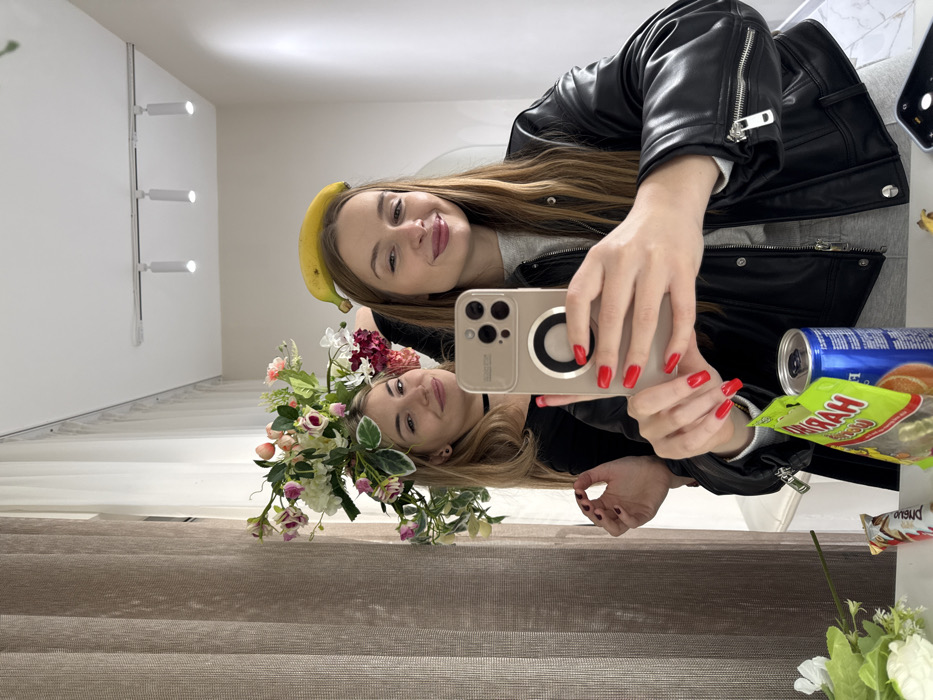 The image depicts a mirror selfie of two women within an indoor setting in Budva, Montenegro. In the foreground, a woman with long brown hair, wearing a black leather jacket over a grey top, holds a gold-colored smartphone horizontally with her right hand, showcasing red nail polish. A yellow banana is balanced horizontally on her head. Her lips are slightly parted in a subtle smile. Reflected behind her, a second woman with long blonde hair wears a black top and a black choker necklace. A large artificial floral arrangement composed of pink, white, and red flowers and green foliage rests on her head. She also smiles towards the camera.
The immediate foreground displays a white countertop holding a green and yellow "HARIBO" branded package, a blue and silver beverage can, and a white and red "Kinder Bueno" chocolate bar wrapper. A second, black smartphone lies screen-down on the right side of the countertop. The background reveals light brown, vertically textured curtains on the left, with sheer white curtains visible behind them. The room features white walls and a white ceiling equipped with a track lighting system containing three white spotlight fixtures.