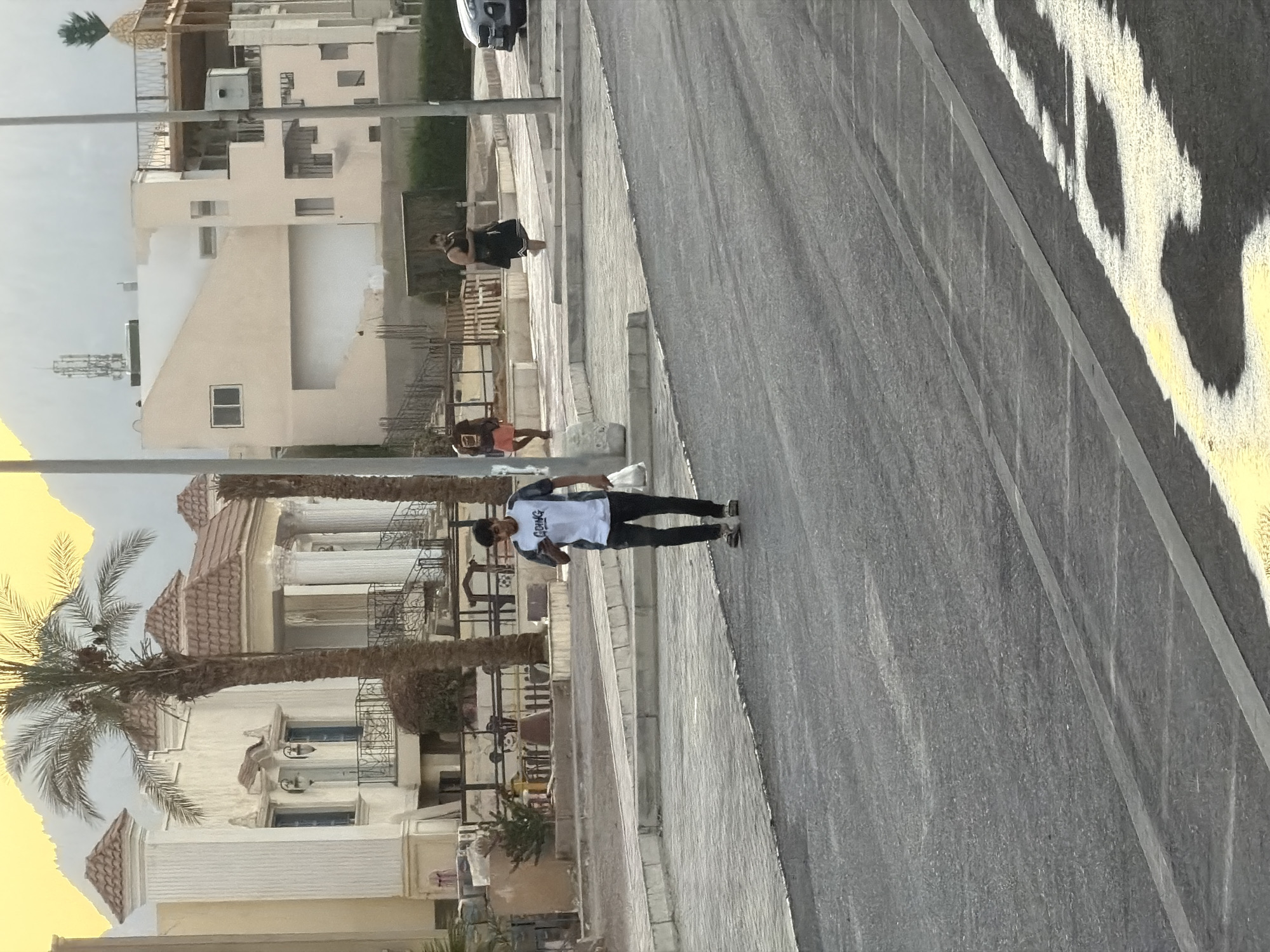 The image depicts a street scene in Sharm El Sheikh, Egypt, with a focus on the architecture and the people present. In the foreground, a young man wearing a white t-shirt with black lettering and black pants walks on the paved road, holding a white plastic bag in his right hand and looking at his phone. Behind him, across a sidewalk, are several buildings with distinct architectural styles. To the left, a large, ornate building with white pillars and decorative roofing is visible, flanked by palm trees. To the right, a lighter-colored building with multiple levels and balconies is seen, topped with a large decorative pineapple. The background reveals mountains. Several other individuals are present in the scene, including a person in red shorts walking further down the sidewalk and another person in a black dress. The lighting suggests it is daytime, possibly late afternoon or early evening, given the warm tones in the sky. The street appears to be under construction or recently paved in some sections, as indicated by the fresh asphalt and yellow markings.