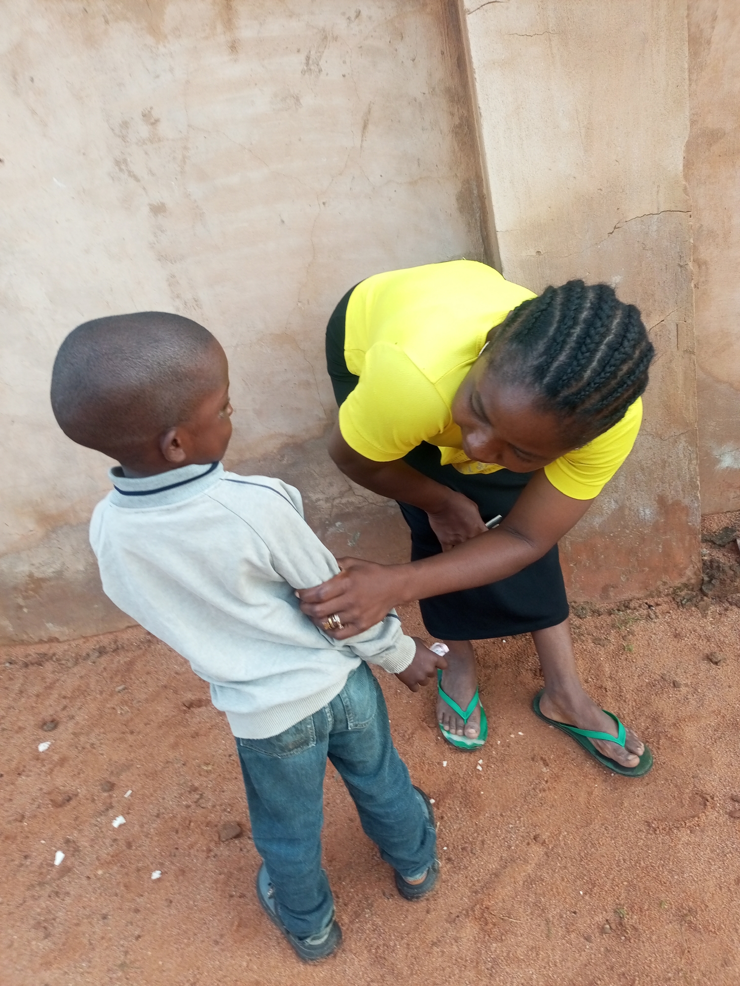 A woman in a bright yellow shirt and a black skirt is bending down to interact with a young boy. The boy is wearing a light gray long-sleeved shirt and blue jeans. The woman has her hair styled in braids and is wearing green flip-flops. The boy is wearing dark-colored shoes. They are standing on reddish-brown dirt, possibly in a rural or outdoor setting. Behind them is a rough, textured wall, likely made of stucco or concrete, with some cracks and discoloration. The scene appears to be outdoors, as indicated by the natural lighting and the ground surface. The interaction suggests a moment of attention or instruction from the woman to the boy, or perhaps a playful exchange. The overall atmosphere is casual and ordinary. The time of day is not explicitly indicated, but the lighting suggests it could be daytime. There is no visible text in the image. The location context is Tempure, Nigeria.