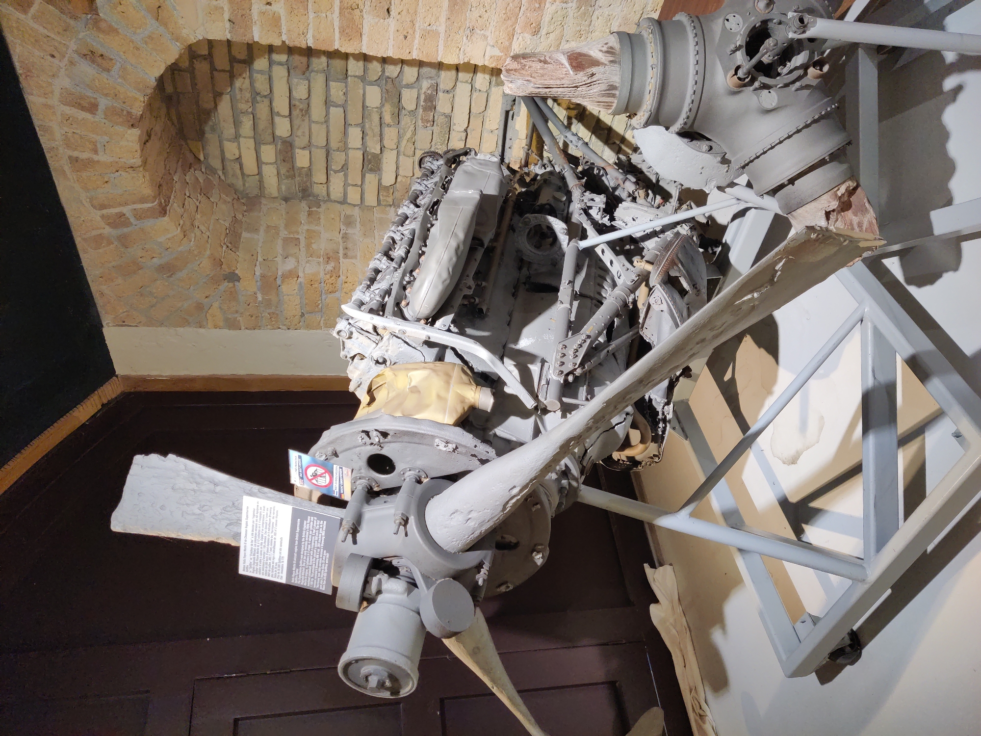 An aviation engine with a propeller is displayed on a stand in a museum, likely in Dunkirk, France, given the context of the Battle of Dunkirk. The engine is gray and appears to be old, with visible mechanical components and a damaged propeller. A informational plaque is attached to the engine, detailing its history and specifications. The background shows a brick wall with an arched opening and a dark wooden door. The floor is light gray, and shadows are cast by the engine and its stand.