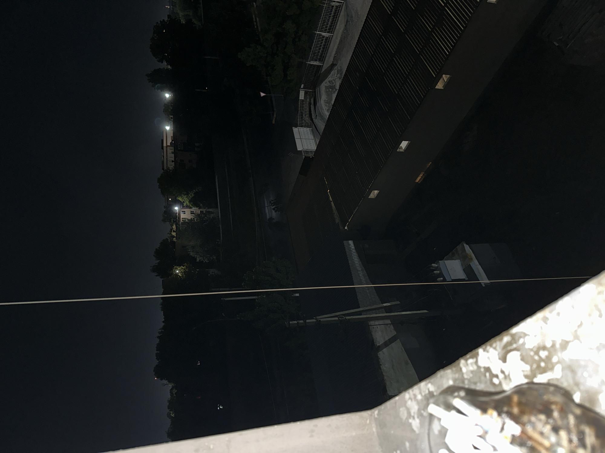 The image shows a nighttime view of an urban area in Fergana City, Uzbekistan. In the foreground, a close-up of an ashtray filled with cigarette butts is visible on a concrete ledge. Beyond the ledge, the scene opens up to a view of buildings and infrastructure. A tall, thin pole, possibly a utility pole, stands prominently in the middle ground. To the right, a long, dark-colored building with a corrugated metal roof is visible. Further in the background, there are several other buildings, some with illuminated windows, suggesting residential or commercial activity. Trees and greenery are scattered throughout the landscape, silhouetted against the dark night sky. Distant lights twinkle, indicating the presence of more urban development. The overall atmosphere is quiet and nocturnal.
