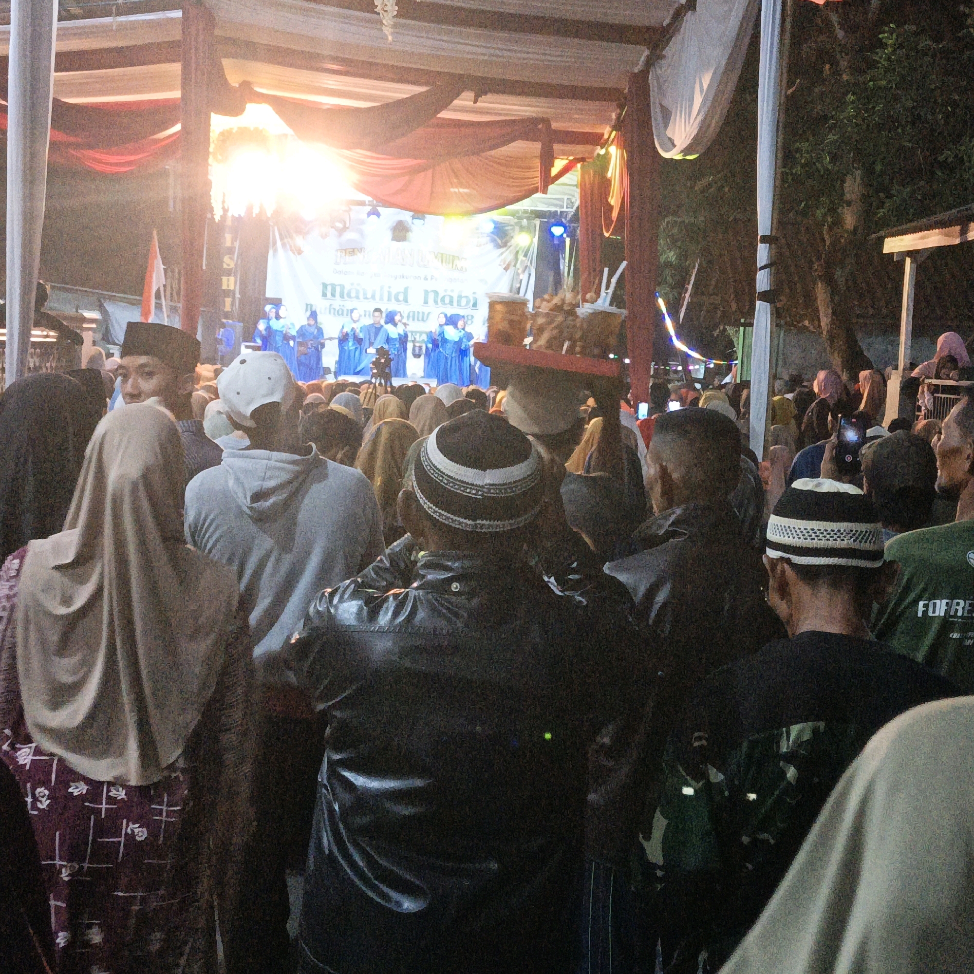 In the city of Kalitorong, Indonesia, a Maulid Nabi celebration is underway outdoors. A crowd of people gathers around a stage, where performers in blue attire are visible. The stage is adorned with a banner that reads "Maulid Nabi" and other Indonesian text, illuminated by bright lights. The event appears to be taking place at night, as indicated by the artificial lighting and the dark sky. The audience consists of men, women, and children, many of whom are wearing traditional Islamic clothing such as peci hats and hijabs. Some individuals are holding up their mobile phones, likely to capture the event. The scene depicts a community gathering celebrating a significant religious occasion.