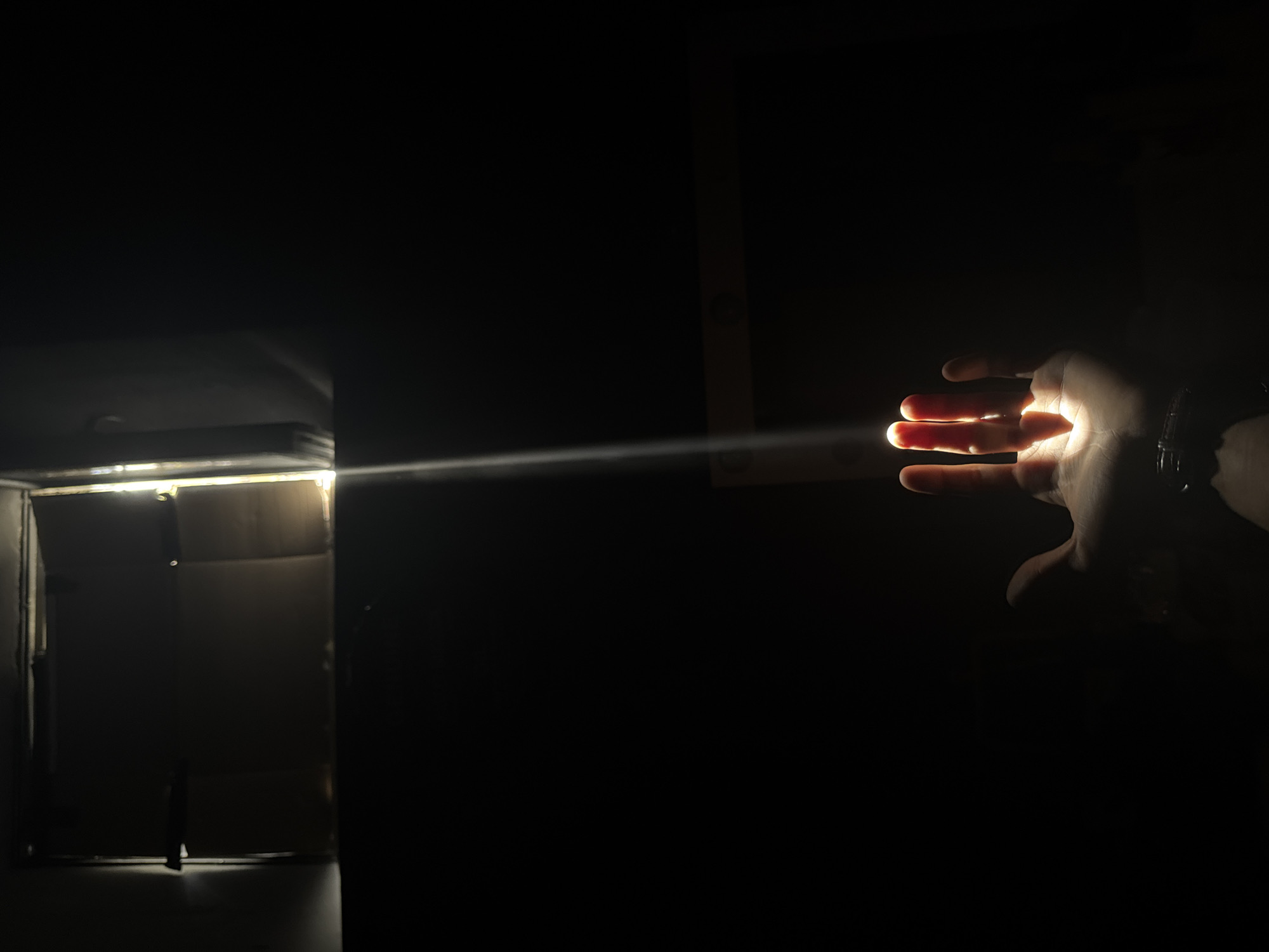 In a darkened space, a horizontal beam of light emanates from a partially open cardboard box on the left towards a hand on the right. The hand is positioned in the path of the light, causing it to illuminate the palm and fingers. The environment is predominantly dark, with a hint of a rectangular structure visible behind the hand. The image does not contain information to verify the location of Moscow, Russia.