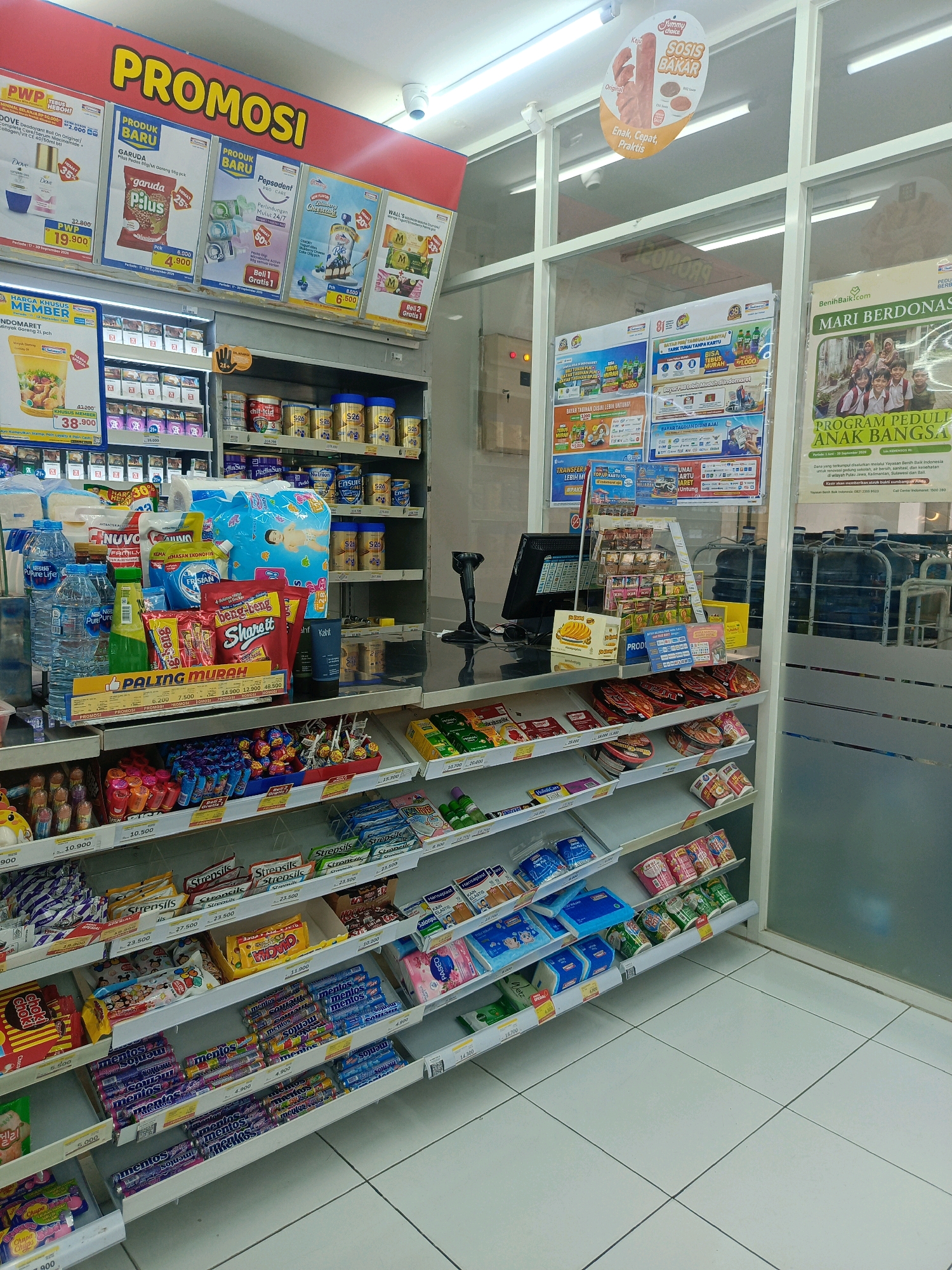 The image depicts the interior of a convenience store, likely Indomaret, in Kepolorejo, Indonesia. The store is well-stocked with various products displayed on shelves. Promotional banners are prominently visible, with a large red banner at the top reading "PROMOSI" (Promotion). Various product advertisements and special offers are displayed on the walls and hanging from the ceiling.
In the foreground, shelves are filled with packaged goods, including snacks, drinks, toiletries, and baby products. Brands like Nuvo, Frisian Flag, Pepsodent, and S26 are identifiable. Candy and lollipops are arranged in bins on a lower shelf. A checkout counter with a computer monitor and barcode scanner is visible on the right side of the image. In the background, large water dispensers are stored.
The store appears to be clean and well-lit, with white tiled flooring. The overall impression is that of a typical Indonesian convenience store operating during business hours, catering to local consumer needs. No individuals are clearly visible in the image, suggesting a moment when the store might be less crowded or captured from a perspective that avoids human presence.