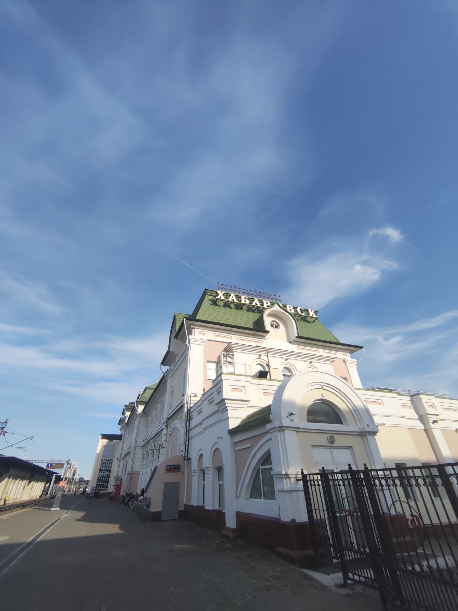 The image shows the Volochaevka-1 railway station building, located in Volochaevka-1, Russia. The building has a light pink facade with white trim and a green roof. The name "ХАБАРОВСК" is visible in large letters on the roof. The sky is blue with some scattered clouds. In the foreground, there is a paved area with a black metal fence. Railway tracks and a platform are visible to the left of the building. Several people are seen sitting on a bench near the building. The scene appears to be a sunny day.