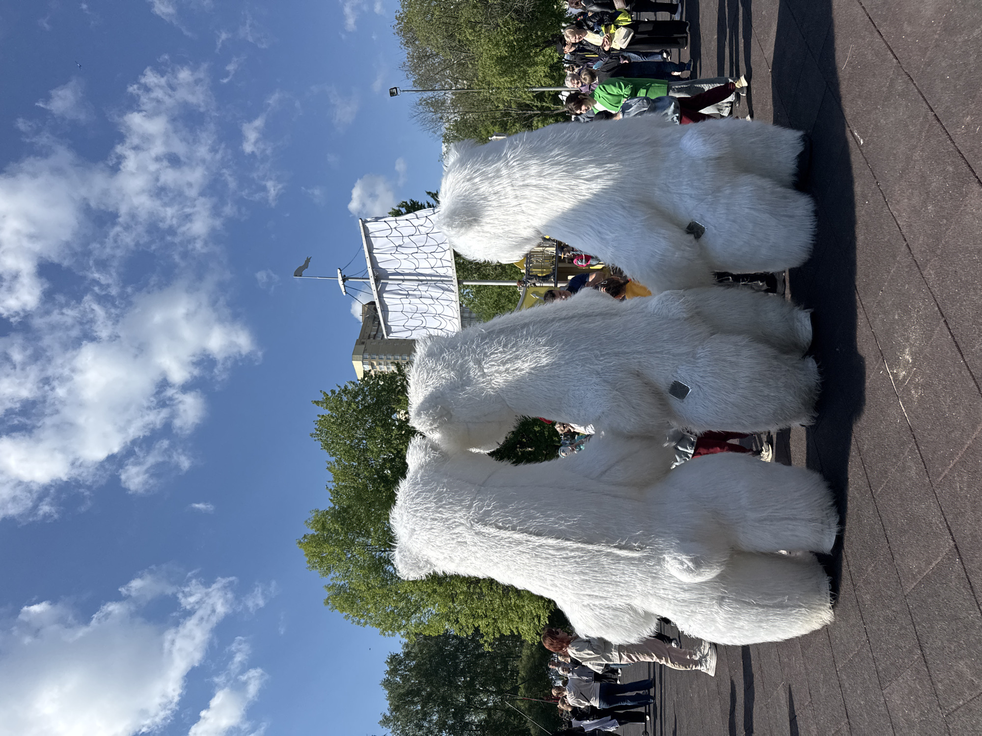Three people wearing large, white, furry costumes resembling polar bears stand on a paved surface, likely a street or square. A crowd of people is visible in the background. Behind the “polar bears” is a structure resembling a ship mast. The sky is blue with scattered clouds. The location is not clearly discernible to be Vsevolozhsk, Russia.