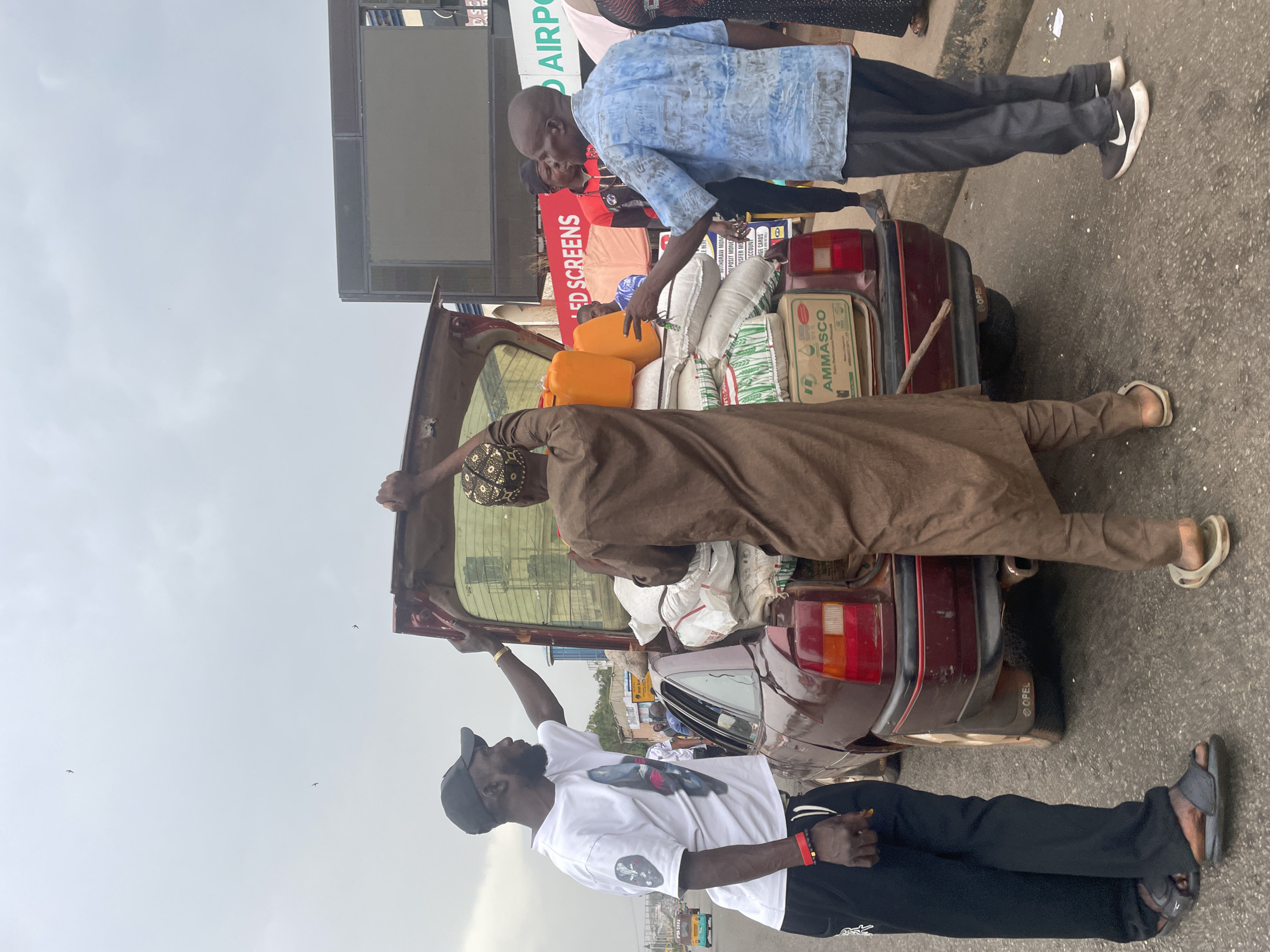 The image depicts a group of three men loading goods into the back of a maroon Opel sedan. The car's trunk is open and filled with sacks and several orange jerrycans. A man in a brown robe is inside the trunk arranging the items, while a man in a light blue shirt and black pants appears to be handing items to him. A third man, wearing a white t-shirt, black pants, and a black cap, stands at the back of the vehicle. The environment suggests an urban setting, with a sign indicating "AIRPORT" and another sign mentioning "ED SCREENS." A sign with the inscription “AMMASCO” can be seen attached to the car. The location is identified as Jos, Nigeria, based on the brand markings on the goods and vehicle.