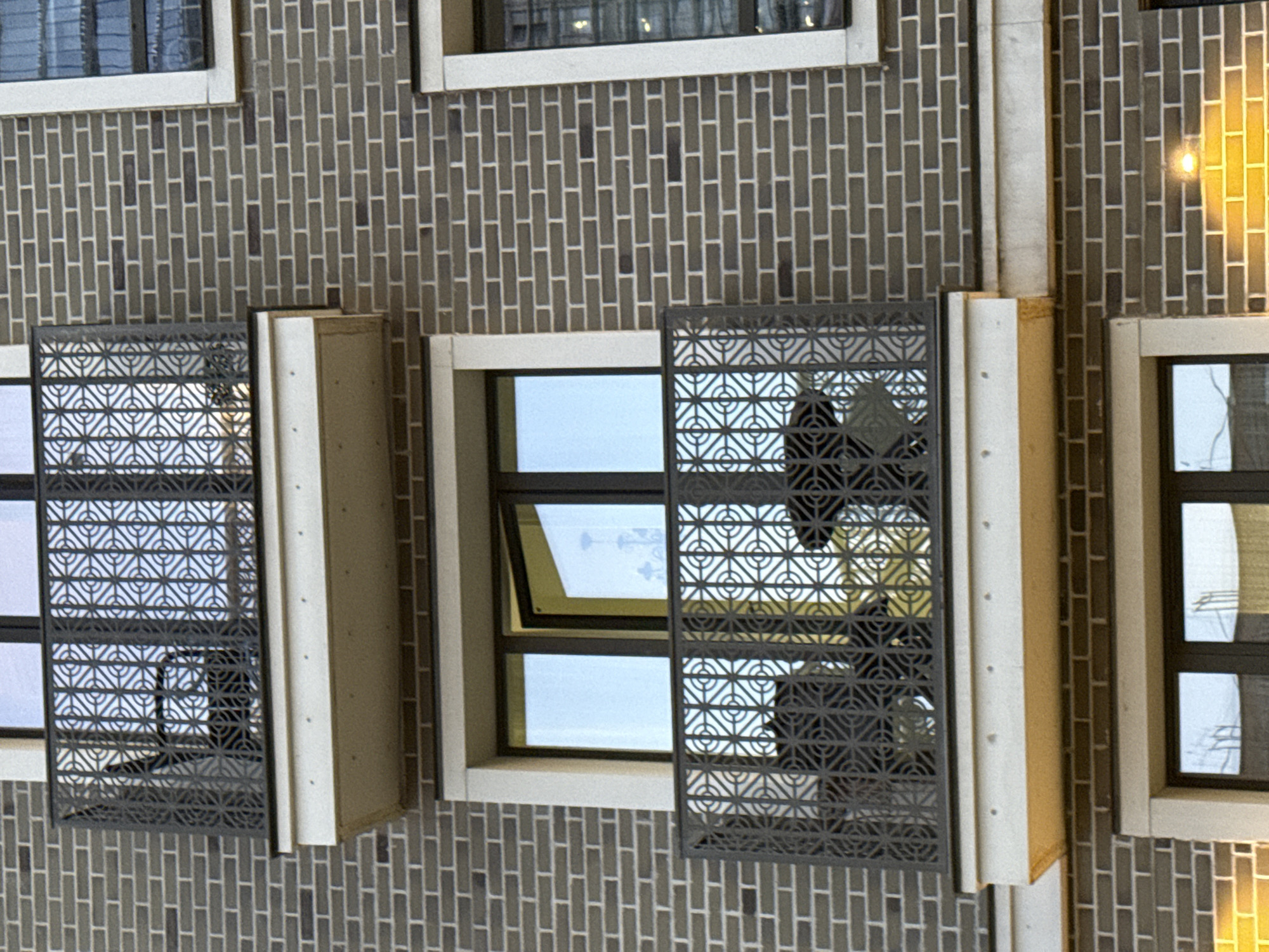 The image shows a section of a building facade, possibly in Moscow, Russia, although there is no explicit evidence within the image to confirm the location. The building is constructed of light gray bricks. Several windows are visible, some of which are partially obscured by decorative metal screens with geometric patterns. One window is slightly ajar. The windows are framed with off-white or beige trim. One window reflects a light source, possibly from a streetlamp or interior light. The scene is well-lit, and there are no people visible in the image.