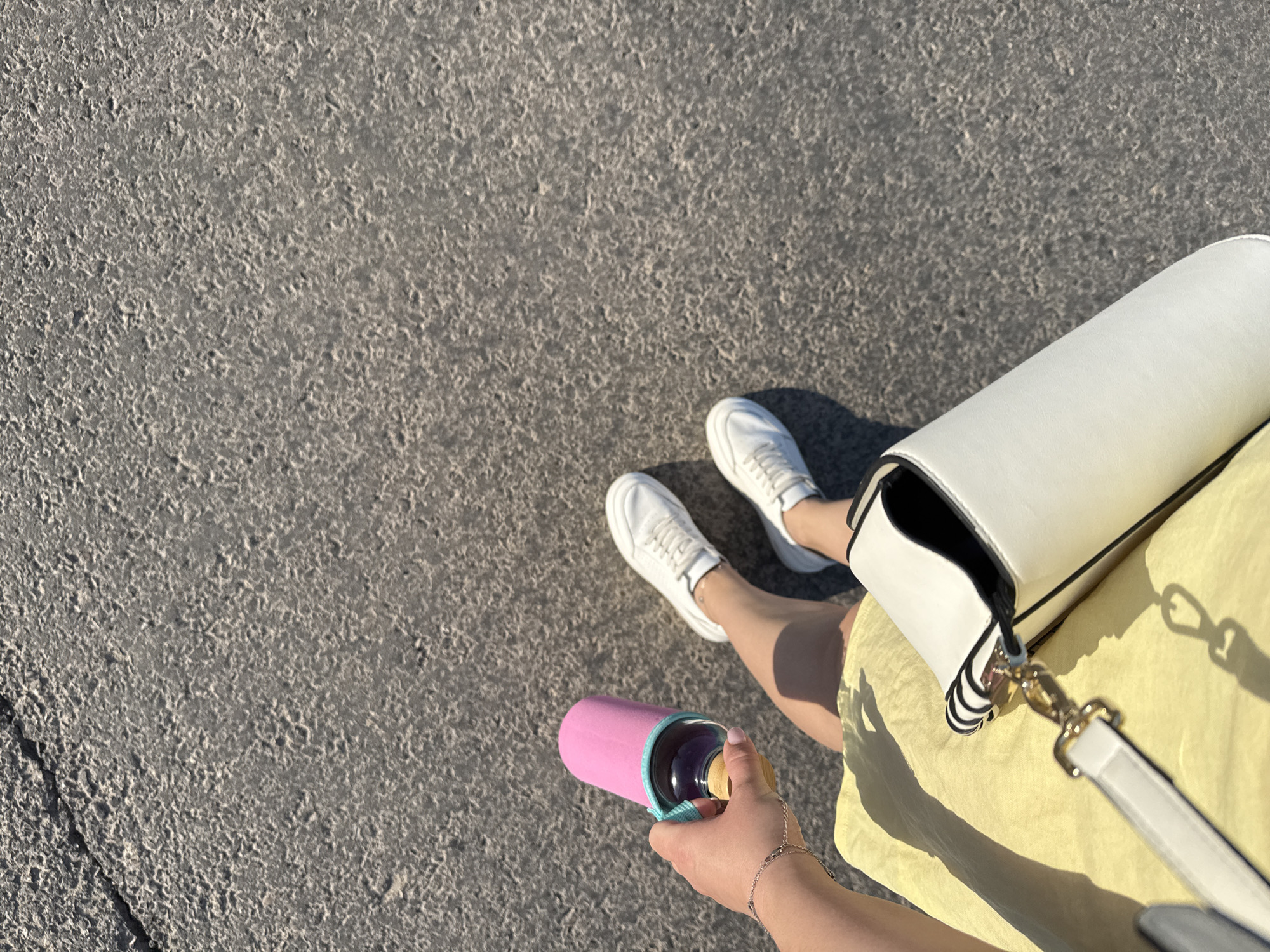 From an overhead perspective, the image shows a person standing on what appears to be an asphalt surface. The person is wearing white sneakers and a yellow dress. They are holding a pink-sleeved bottle in their hand. Beside the person, there is a white purse with a shoulder strap. The image does not provide sufficient information to determine if the location is Izhevsk, Russia, or to identify specific actions, interactions, or other details of the scene.