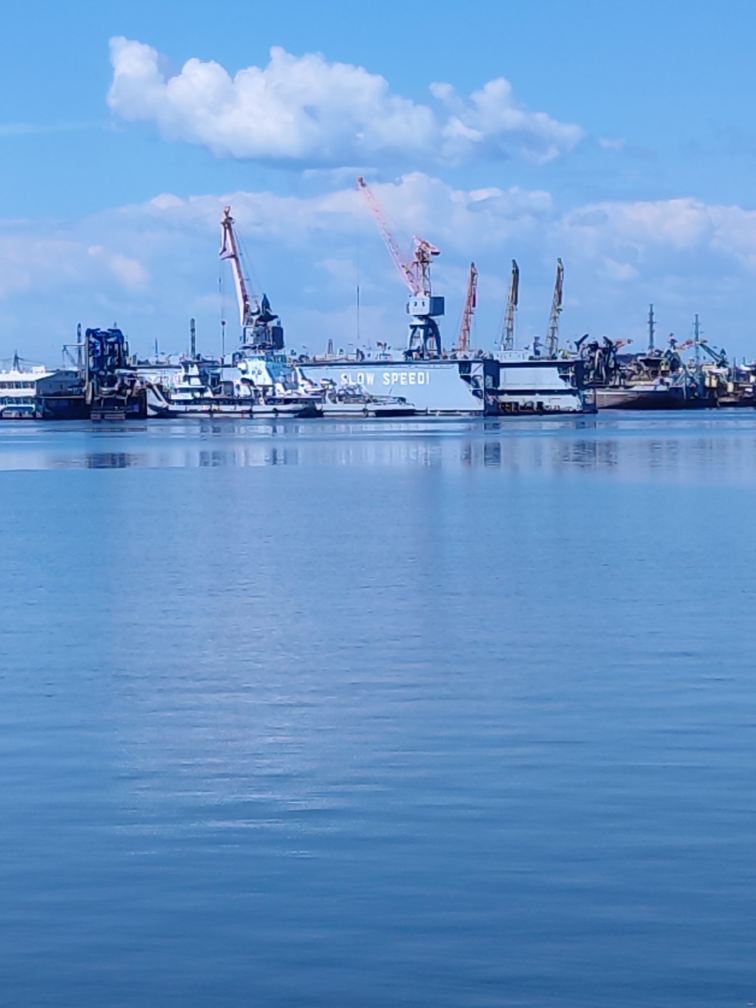 A wide shot captures a blue sky with scattered white clouds above a calm, blue body of water. Along the horizon, a port or shipyard is visible, characterized by a collection of industrial structures and vessels. Several large cranes, some with pink booms, dominate the skyline, suggesting ongoing construction or repair work. A long, blue vessel with white lettering reading "SLOW SPEED" is prominently featured. Other ships and structures, including what appears to be a passenger ferry on the far left, are also present in the background, contributing to the busy industrial atmosphere. The water reflects the sky and the structures above, creating a serene contrast with the industrial activity. The scene is likely located in the city of Chaikovsky, Russia, a city known for its port on the Kama River and its industrial sector. There are no people visible in the image.