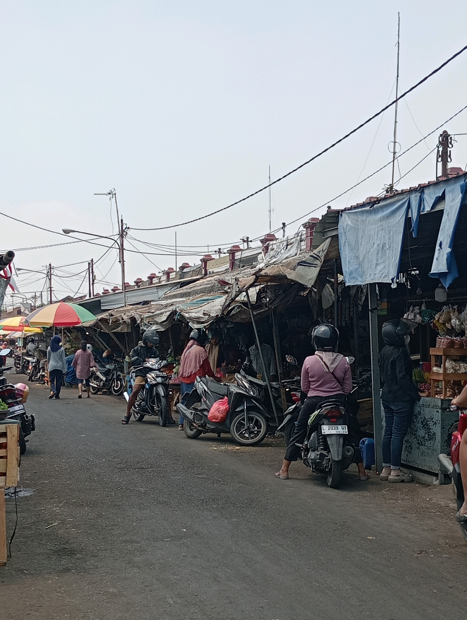 The image depicts an outdoor market scene in Jagalan, Indonesia. A paved road runs through the market, lined with stalls constructed from makeshift materials like tarps and wood. Several motorcycles are parked along the road, some with riders. People are visible browsing and interacting within the market. Colorful umbrellas provide shade over some of the stalls. The overall atmosphere suggests a bustling local marketplace.