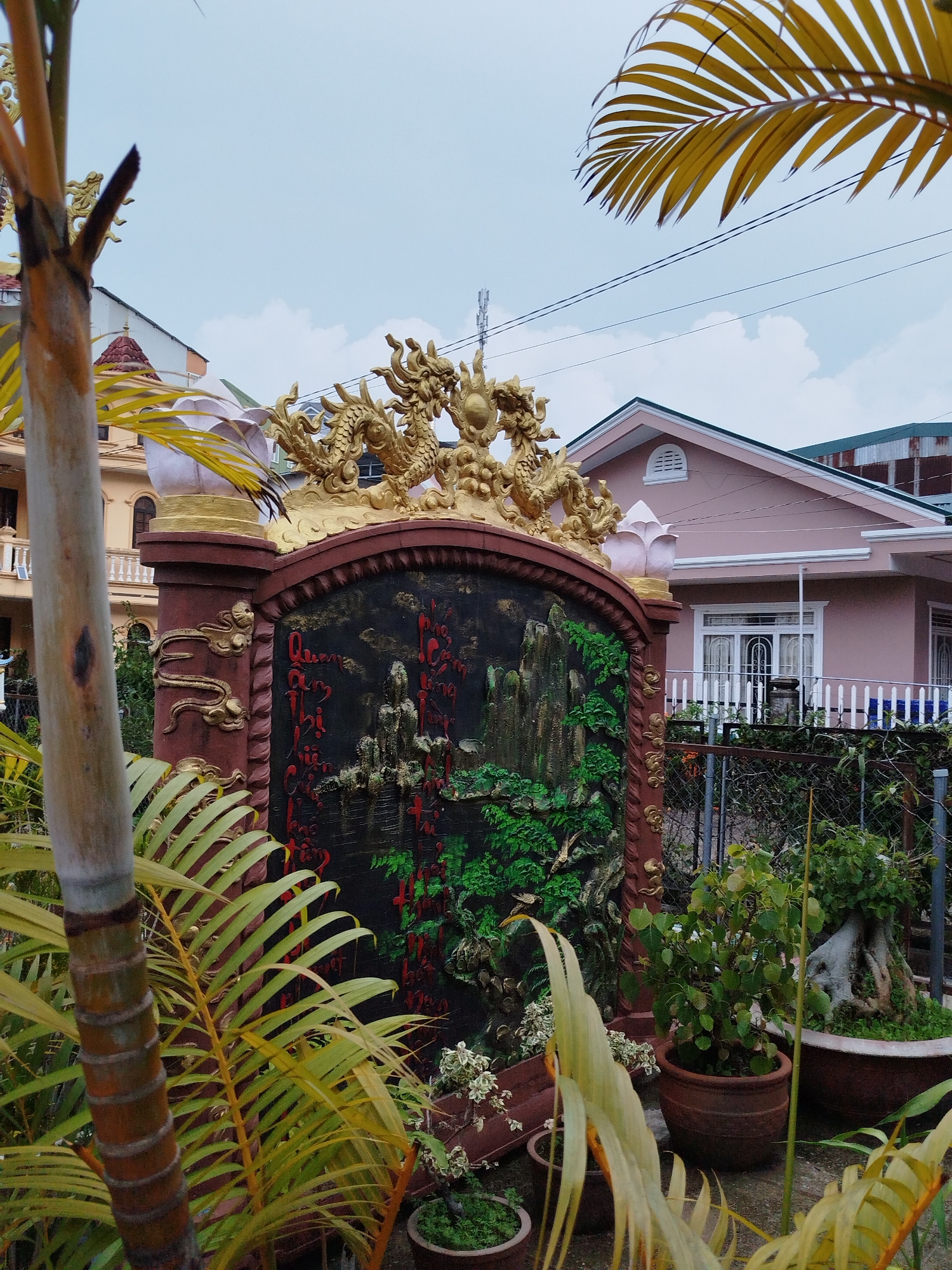 The image captures a vibrant garden scene in Xuân Hương - Đà Lạt, Vietnam. A decorative structure stands as a focal point, featuring a black panel adorned with raised imagery of mountains and greenery, topped with intricate golden dragon sculptures. The structure is framed by brown pillars with golden embellishments. Red Vietnamese calligraphy is visible on the black panel.
The foreground is filled with a variety of potted plants with green and yellow foliage, partially obscuring the base of the decorative structure. Behind the garden, a pink house with a white picket fence can be seen. Overhead power lines are visible against the overcast sky.