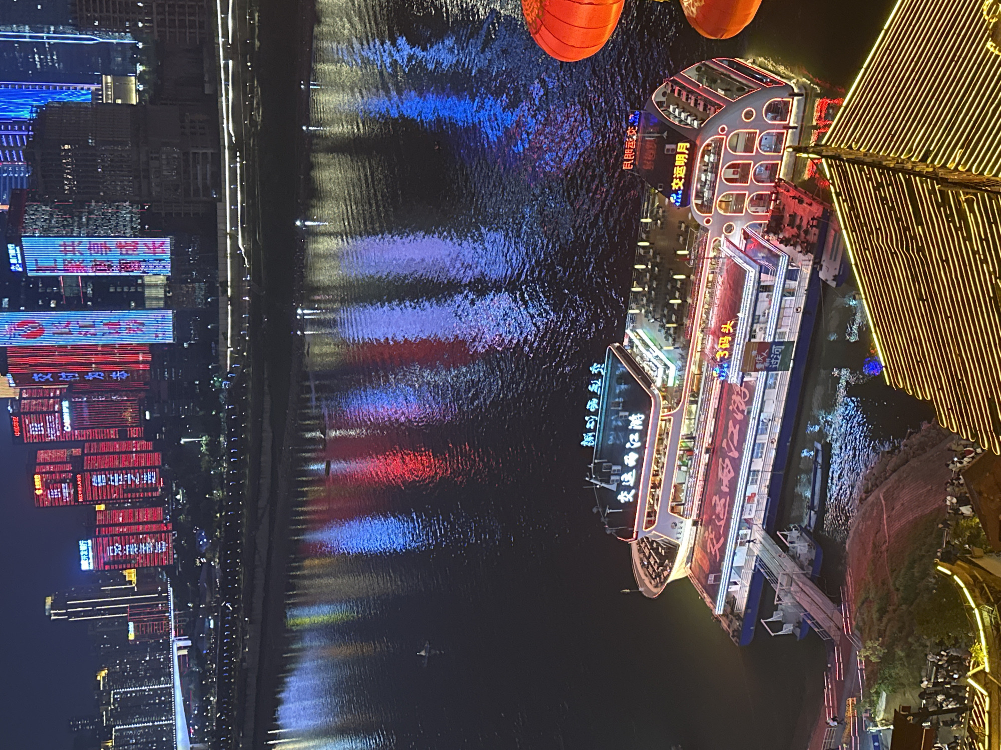 Here is a description of the image:
The image showcases a vibrant night scene in Yuzhong District, Chongqing, China. A body of water reflects the colorful lights of the city skyline and illuminated boats. On the left, tall buildings display bright, animated advertisements and patterns. Across the water, a large passenger boat is docked, adorned with glowing lights and Chinese characters. Red lanterns hang above the boat, adding to the festive atmosphere. To the right, a structure with dense, yellow lighting overlooks the water. Below, there is a crowd of people along the riverbank.