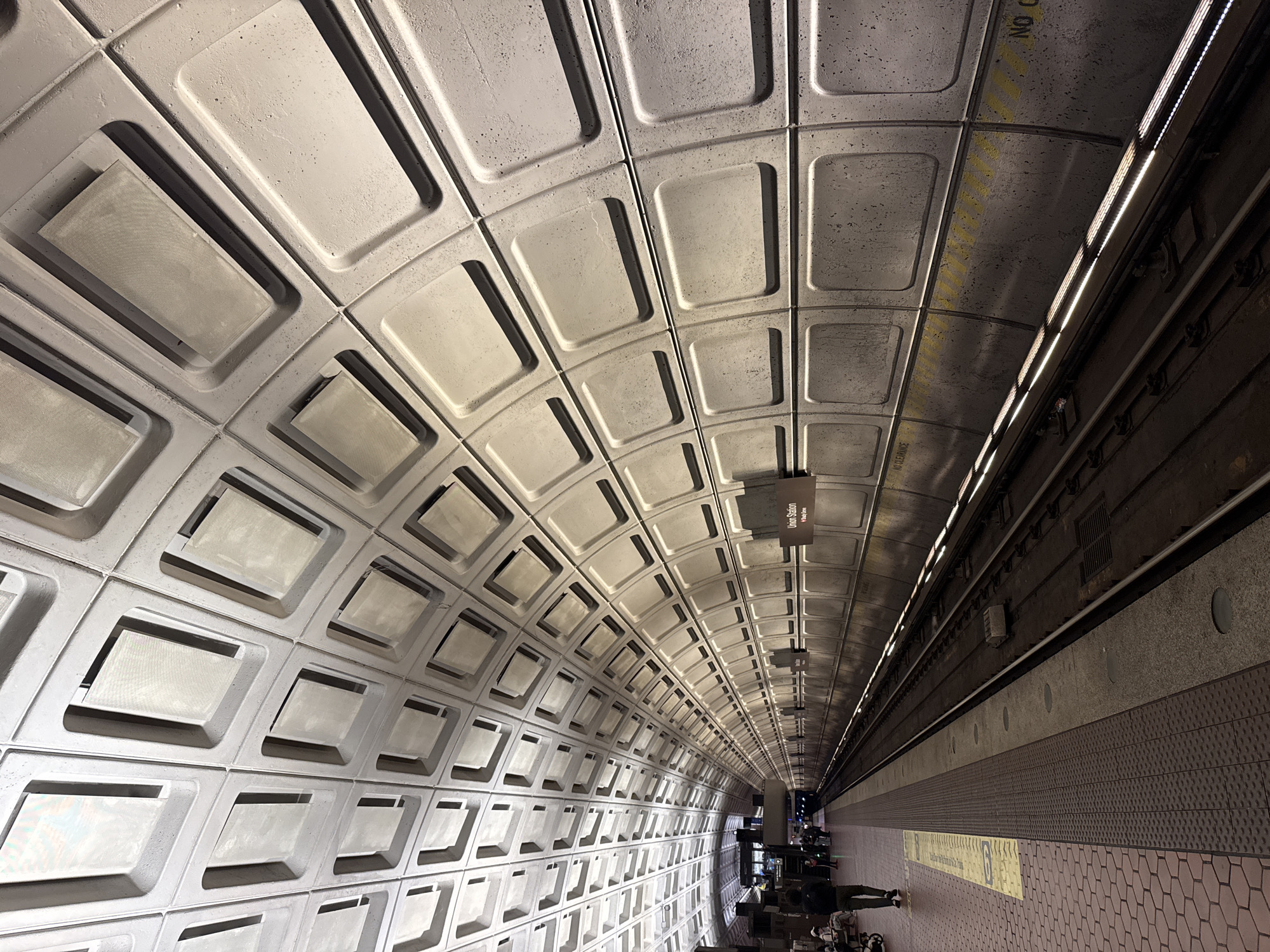 This image presents a striking view of an underground metro station, identified by a hanging sign as "Union Station." The camera is angled upwards and along the length of the station, capturing the iconic, vaulted concrete ceiling that dominates the frame.
The ceiling is composed of numerous coffered panels, primarily light gray, with a textured, almost rough finish. Many of these recessed panels contain rectangular, mesh-covered fixtures, likely housing lighting elements or ventilation systems. The vaulting creates a strong sense of depth and architectural grandeur, characteristic of the Washington Metro system.
On the right side of the image, the metro platform extends into the distance, paralleling the tracks. Several individuals are visible on the platform; some are seated on benches, while others stand, presumably waiting for a train. The platform floor features a distinctive pattern of reddish-brown hexagonal tiles and a darker, textured section closer to the tracks. A prominent yellow tactile warning strip runs along the platform edge. Beyond this, the dark tracks and associated power infrastructure are visible, illuminated by a continuous strip of bright artificial lights embedded in the tunnel wall.
A black rectangular sign hangs above the platform, clearly displaying "Union Station" in white letters, with smaller text below it, likely indicating the metro line. On the ceiling above the tracks, a yellow warning stripe is visible, with partially legible text that includes "NO CLEARANCE." The station is brightly lit by artificial light, suggesting an indoor setting where natural light is absent. The scene captures the typical ambiance of a public transit hub, with people awaiting transportation in a distinctly designed urban underground space.