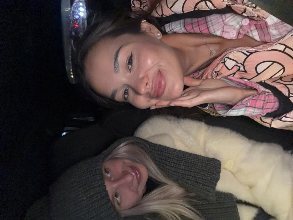 Two women are seated inside a vehicle at night. The woman on the left, with blonde hair, wears a dark grey knit beanie and a matching knit scarf over a light-colored, possibly off-white, faux fur coat. She is positioned slightly to her right, with a faint smile. The woman on the right, with dark brown hair, wears a pink and white patterned collared shirt, a silver necklace with a clover pendant, and a diamond stud earring in her right ear. She is smiling broadly with her right hand placed against her cheek. The background is dark, revealing a blurred urban environment with scattered white and red lights through the vehicle's rear window. The specific location within the city of Krasnoyarsk, Russia, is not discernible from the image.