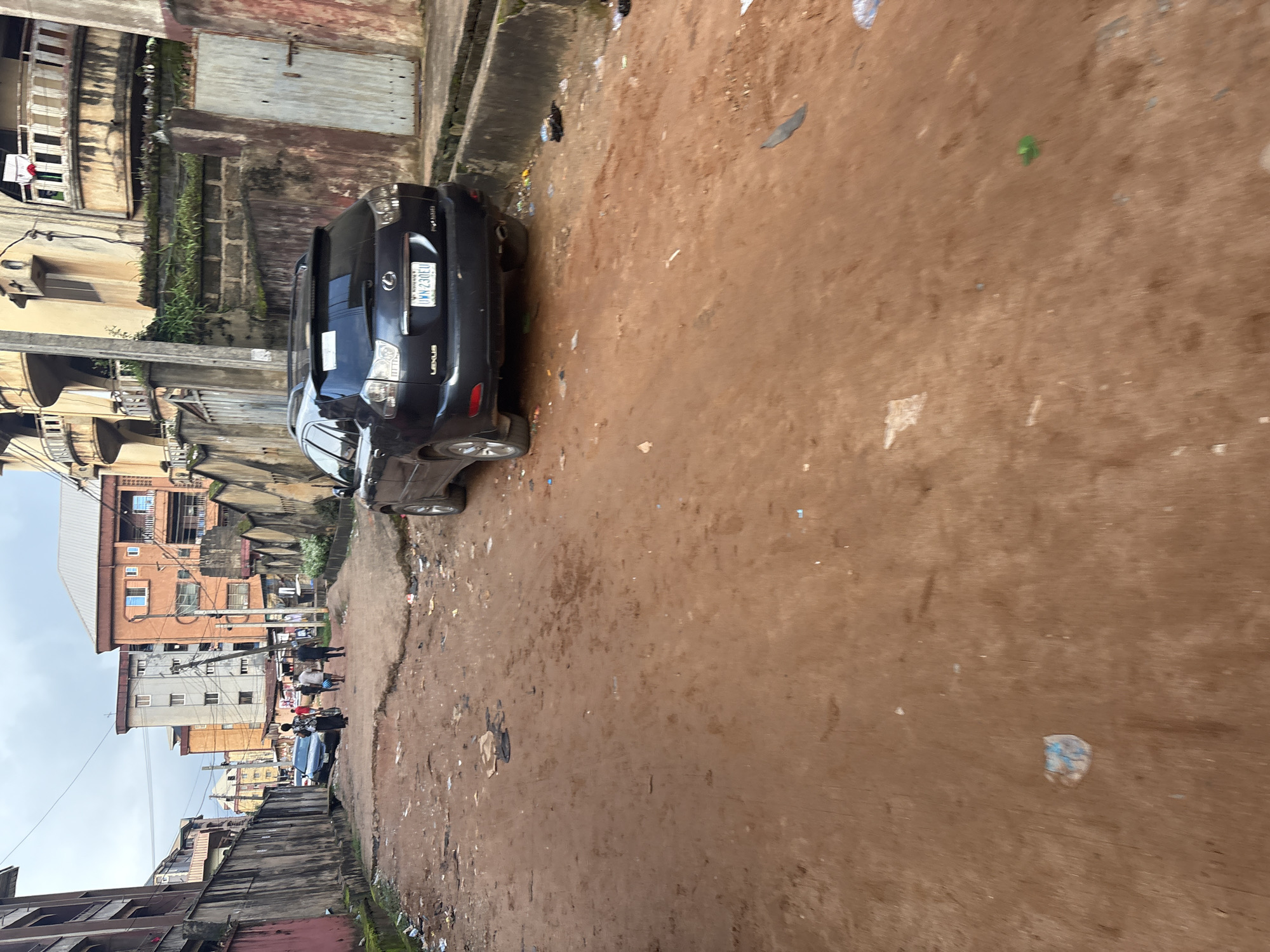 The image depicts a street scene in Onitsha, Nigeria, characterized by a dirt road and surrounding residential buildings. A dark-colored Lexus SUV is parked on the right side of the road. Several people are walking in the distance on the left side of the road. The environment appears somewhat neglected, with debris scattered on the ground. The buildings show signs of wear and tear. The sky is overcast.