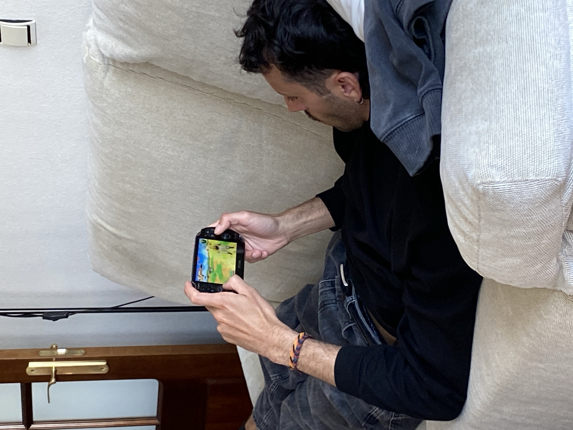 A man sits on a light-colored sofa. He is holding a PlayStation Vita in his hands and appears to be looking at the screen. He is wearing a black long-sleeved shirt and grey jeans. He has a colorful bracelet on his wrist. The background shows part of the sofa, a wooden cabinet, and a white wall. The location in the city of Gijón/Xixón, Spain is unknown.