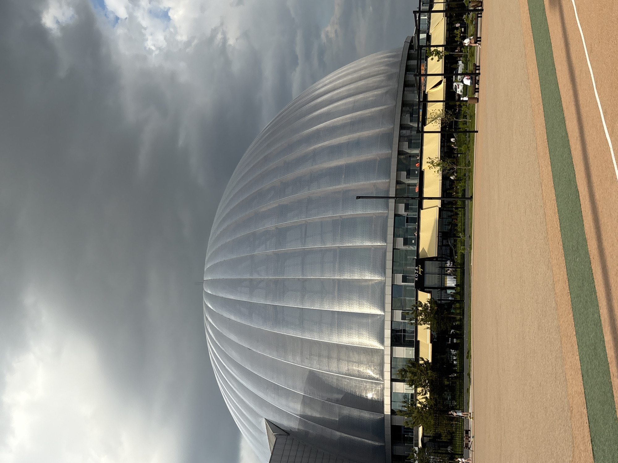 The image displays a prominent, large, modern stadium with a distinctive silver, ribbed, and domed roof, which appears highly reflective under the cloudy sky. This structure, consistent with Gazprom Arena (Krestovsky Stadium), is located in Saint Petersburg, Russia.
Below the main domed section, the building features several stories with extensive glass facades, reflecting the sky and surrounding elements. Attached to this lower section are outdoor areas with yellow awnings and some visible trees and shrubs.
The foreground features a wide, paved outdoor area, possibly a plaza or track, characterized by a light brown or tan surface with a distinct green stripe running lengthwise and several narrower white lines.
The sky above is heavily overcast and grey, with thick clouds dominating the frame, though some lighter patches suggest indirect sunlight or a break in the cloud cover. A few indistinct people are visible in the distance on the paved area and near the awnings, suggesting casual public activity. The overall lighting indicates it is daytime, likely late morning or afternoon under an overcast sky.
A partial text "KOZA" is visible on a sign above one of the glass sections of the building, possibly indicating a shop or cafe. The scene captures a modern urban setting dominated by impressive architecture and a moody sky.