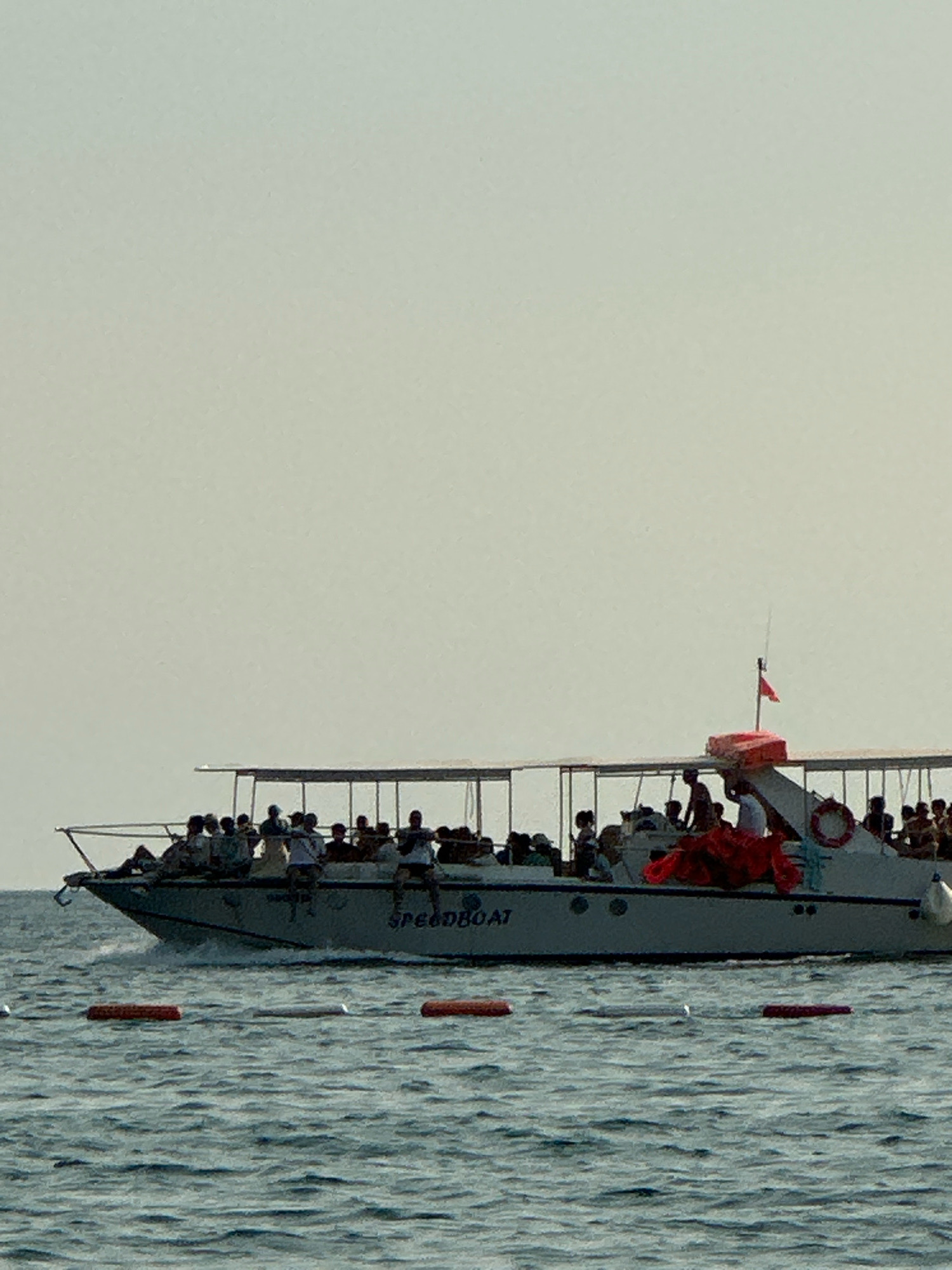 An eye-level, medium shot captures a white speedboat filled with passengers moving across moderately choppy, blue water. The boat has a covered top extending most of its length, supported by poles. Several people are visible standing and sitting on the boat's deck, some appearing to be tourists. The word "SPEEDBOAT" is clearly visible on the side of the vessel. A small red flag flies from a pole near the back of the boat. In the foreground, three orange buoys are floating on the water's surface. The sky is a pale, hazy blue, indicating a bright day. The scene appears to be a typical tourist excursion on the water, likely in a coastal area. Without further information, the specific location in Boreti, Montenegro, cannot be definitively identified from the image alone, but the setting suggests a maritime activity common in such regions.