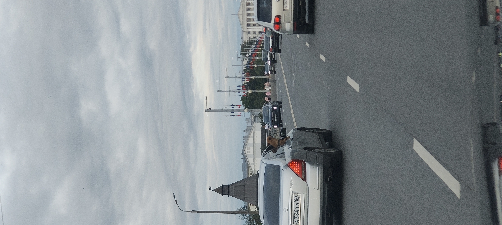 The image captures a view from a vehicle driving on a street in Pskov, Russia, likely on a bridge or elevated road, with a cloudy sky overhead. In the foreground, the rear of a silver Nissan Teana sedan is visible, with a tan dog with its head and upper body out the open rear passenger window. The license plate of the Nissan reads "A334TA 60".
Further down the road, other vehicles are present, including a black SUV and a white SUV. In the background, a line of flagpoles with flags, a lamppost, and buildings can be seen. The architecture of a wooden tower with a dark roof, reminiscent of historical Russian design, is also visible to the left. The road surface is asphalt with white lane markings. The scene suggests a typical urban environment during daytime, possibly with a slight overcast.