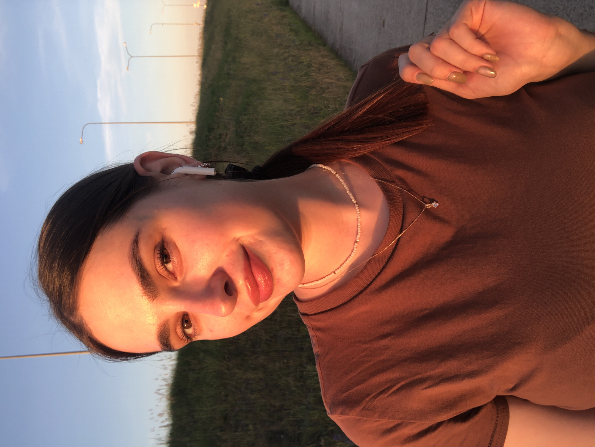 A young woman with dark hair pulled into a ponytail is captured in a selfie taken outdoors. She is wearing a brown t-shirt, a delicate beaded necklace, and a gold necklace with a small pendant. White wireless earbuds are visible in her left ear. Her right hand, with polished nails, is positioned near her neck. The warm, golden light suggests it is either sunrise or sunset. In the background, a grassy embankment slopes upwards, with a paved path and streetlights lining the road. The sky is a clear blue with some wispy clouds. The image appears to be taken in an urban or suburban setting, potentially near a highway or a park, given the presence of streetlights and the overall landscape. The specific location in Noviy, Russia is not discernible from the visual information provided in the image.