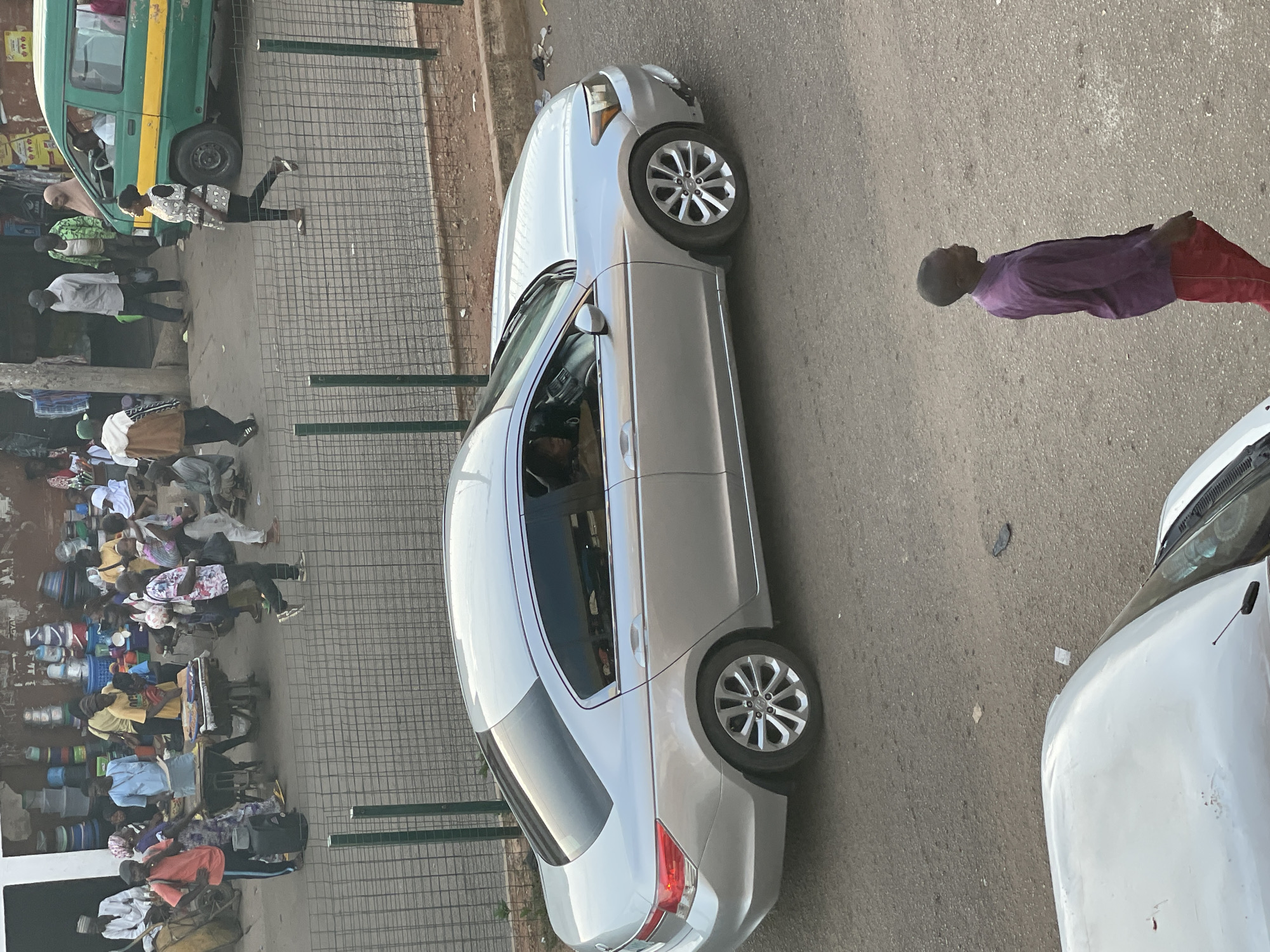 The image shows a street scene in Bukuru, Nigeria. A crowd of people is gathered on the left side of the frame, likely at a market or commercial area. People are dressed in various styles of clothing, and some appear to be carrying bags or goods. A green van and a silver car are parked along the side of the road, next to a wire fence. A man wearing a purple shirt and red pants is walking along the right side of the road. A white car is partially visible in the lower right corner of the image. The setting appears to be a busy urban environment.